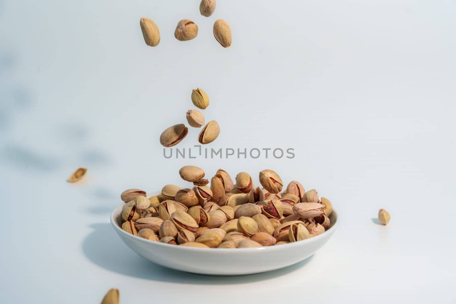 Shelled pistachio nuts captured in motion as they fall into a white bowl already filled with pistachios, against a light-colored background.