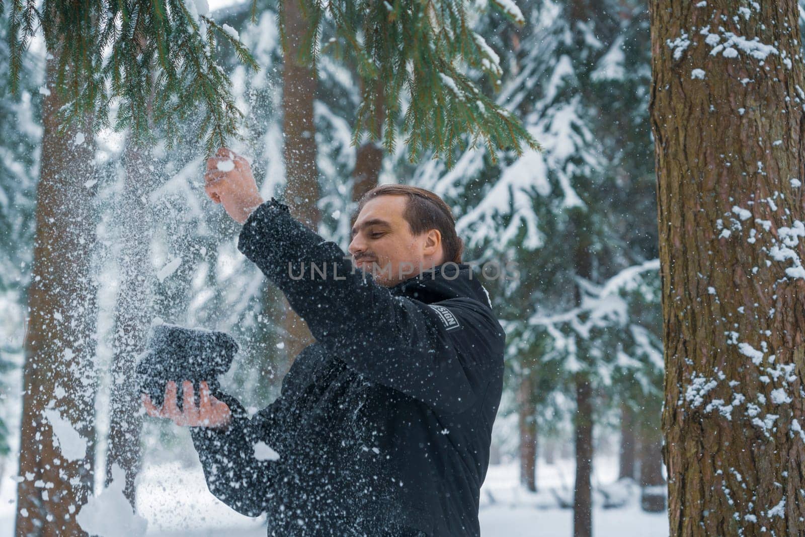 Adult man in black jacket knocking snow from evergreen tree in snowy forest during daytime