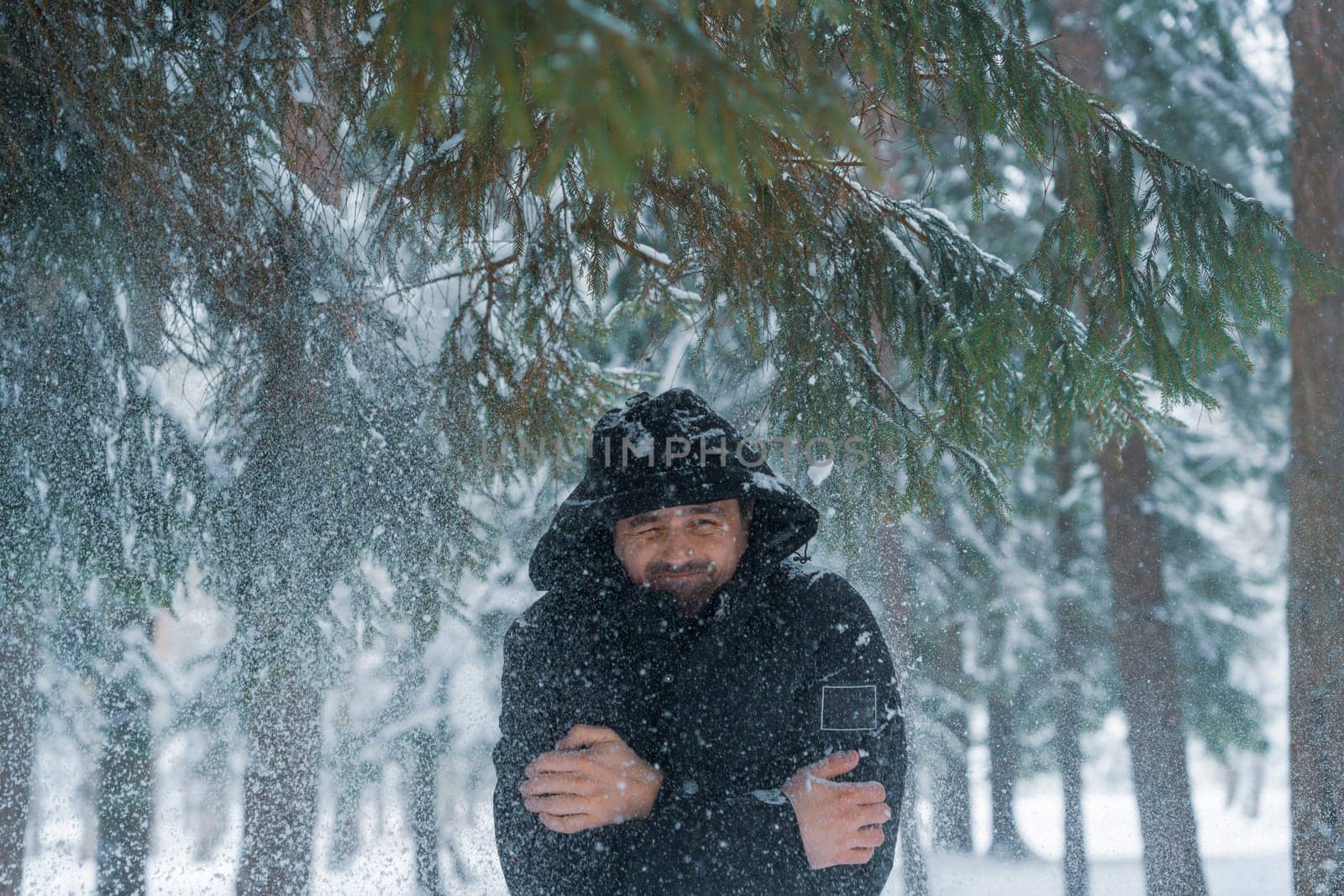 Hooded man in black coat reacts to snow falling from above while standing in snowy forest surrounded by pine trees