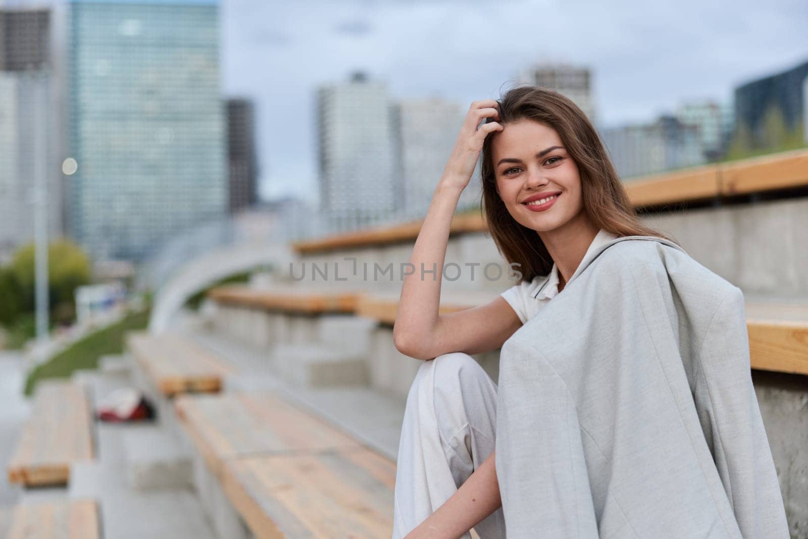 Young woman in casual light outfit sits on outdoor wooden benches smiling confidently with city skyline in background, spring season, fashion lifestyle and relaxation concept