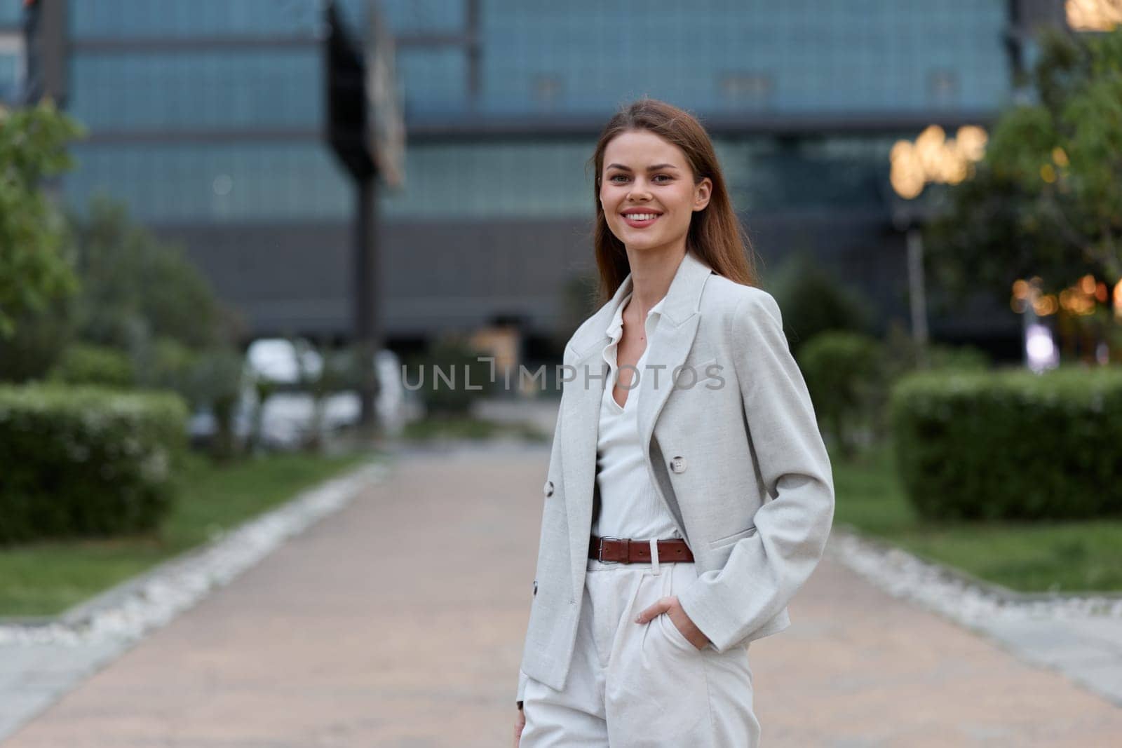 Confident young woman dressed in a light gray business suit stands smiling outdoors in a modern urban park with green foliage, projecting professionalism and modern business style.