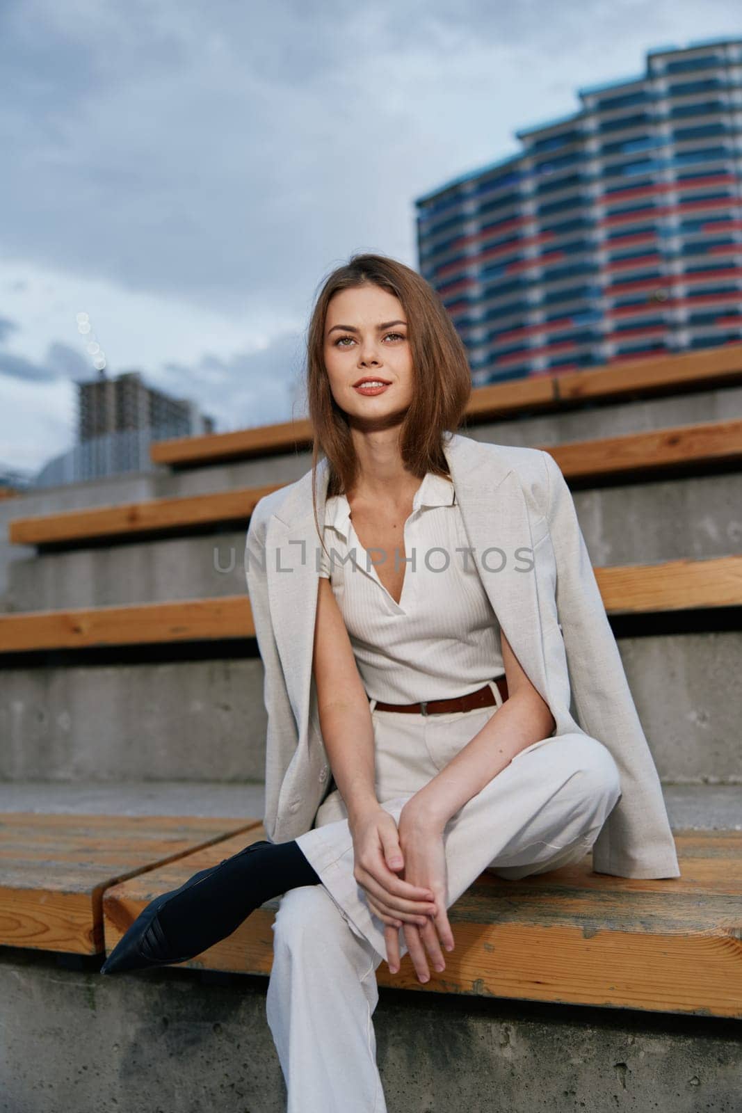 confident woman wearing white business outfit and beige blazer sits on wooden stairs in urban environment with modern architecture and cloudy sky, expressing professionalism and calmness