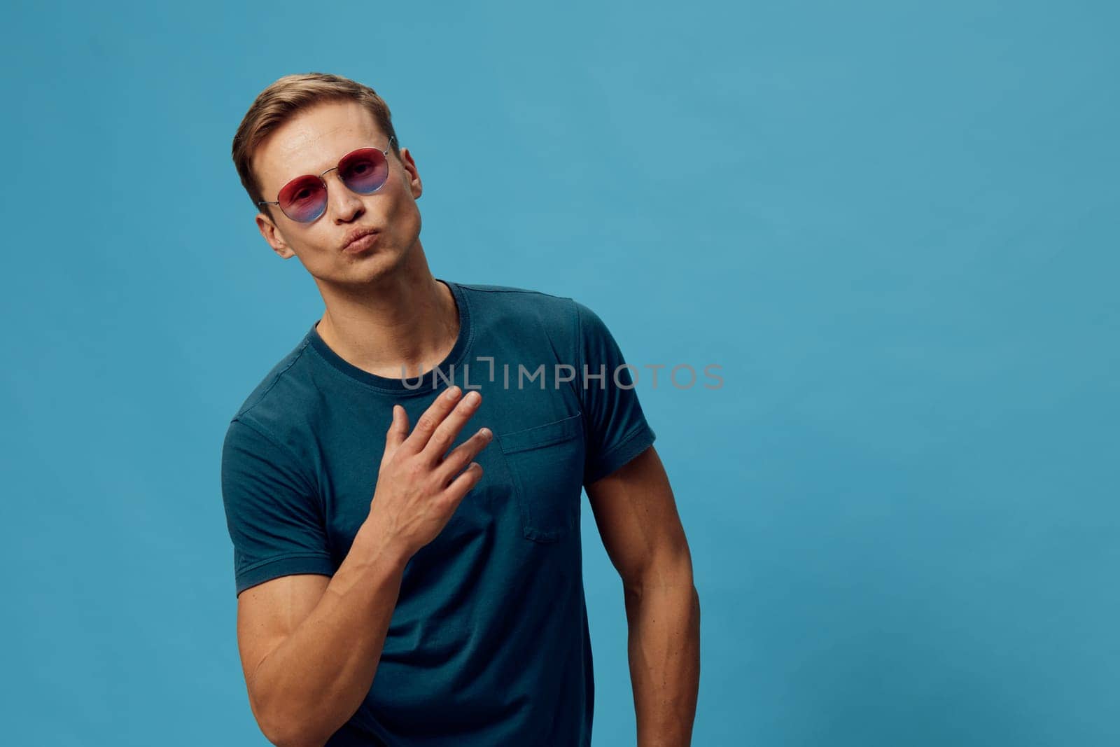 Confident young man with trendy sunglasses and casual blue T-shirt standing in front of a solid light blue backdrop. Male model with cool style, relaxed pose, and fashionable appearance, conveying youthful confidence. Concept: modern fashion.