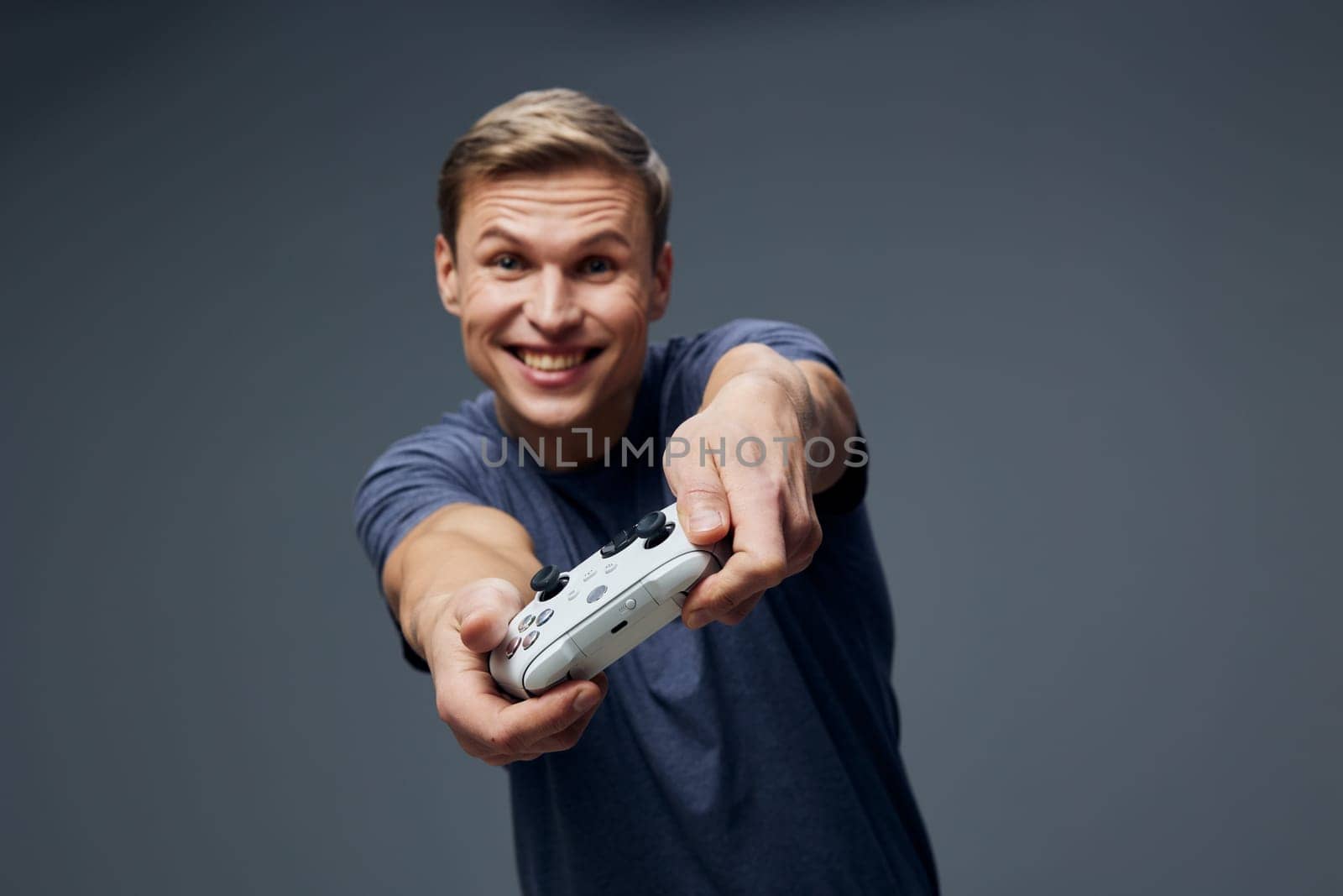 Happy young man with short brown hair and blue casual shirt holding a game controller, smiling enthusiastically while looking at the camera against a plain gray background, capturing excitement and engagement, lifestyle and gaming concept