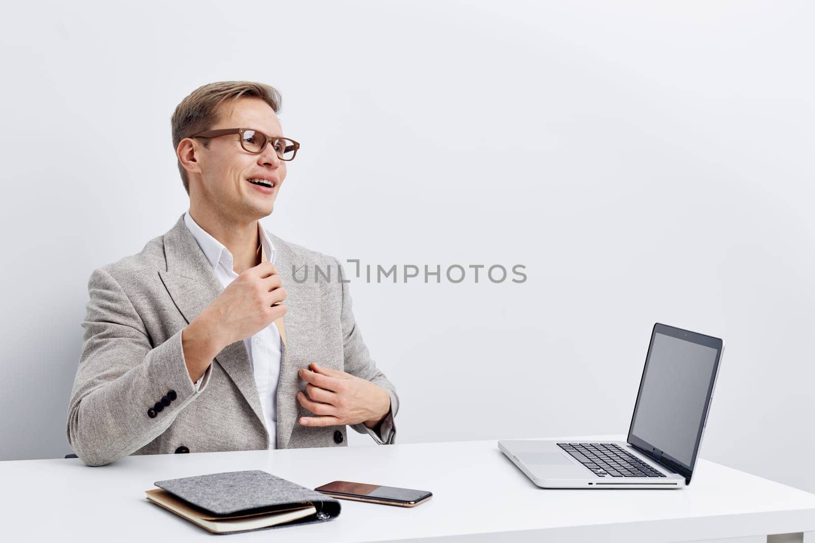 smiling young caucasian man wearing glasses and gray blazer sitting at white desk with laptop, phone, notebook, and remote control, looking aside while talking during video call about working remotely as digital nomad in modern white office. Concept: digital nomad