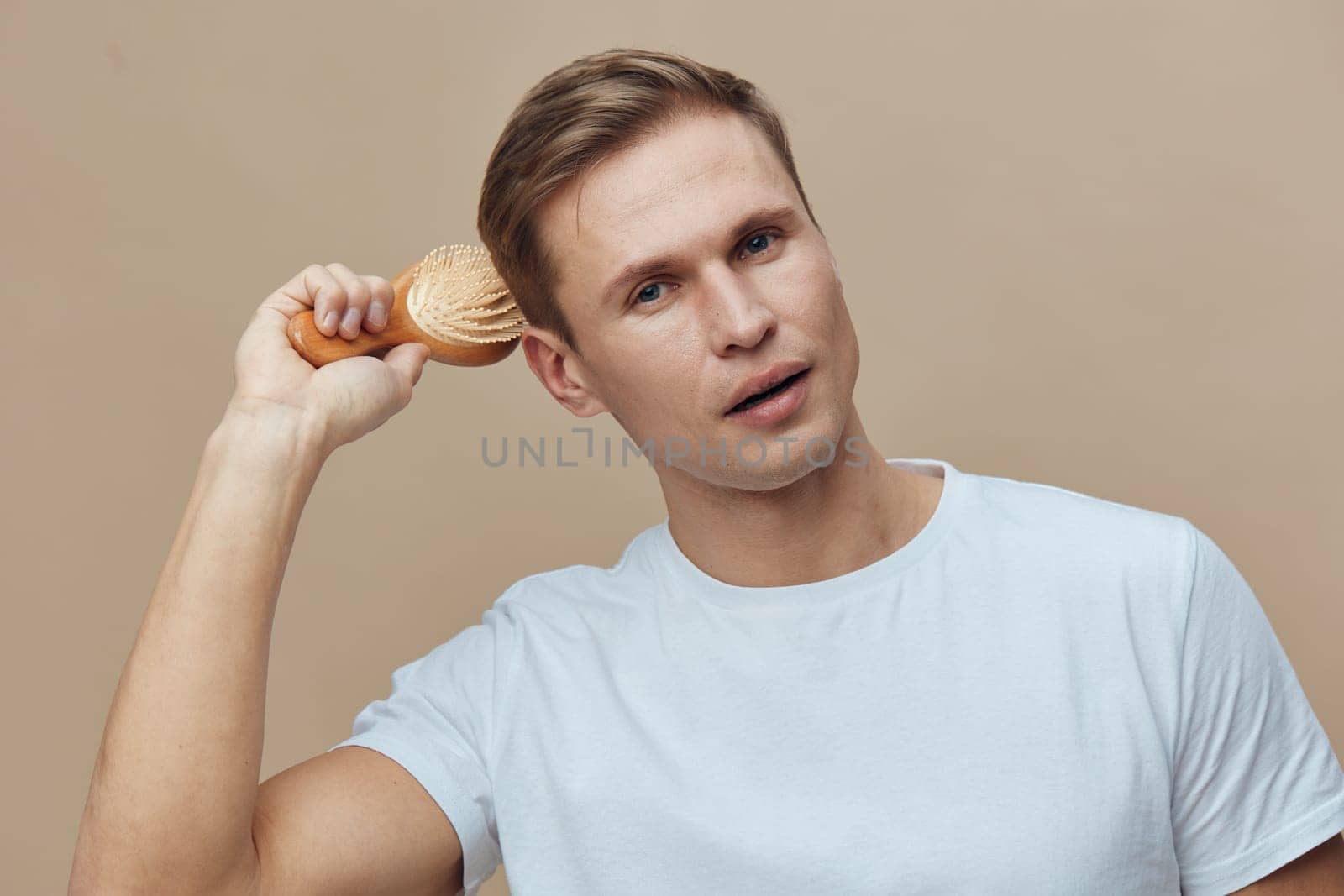 Relaxed young man with light brown hair and blue eyes, dressed in a simple white t-shirt, posing against a beige backdrop, holding a wooden brush near his ear. Studio shot emphasizing grooming, style, personal care, male lifestyle, natural pose, beauty care.