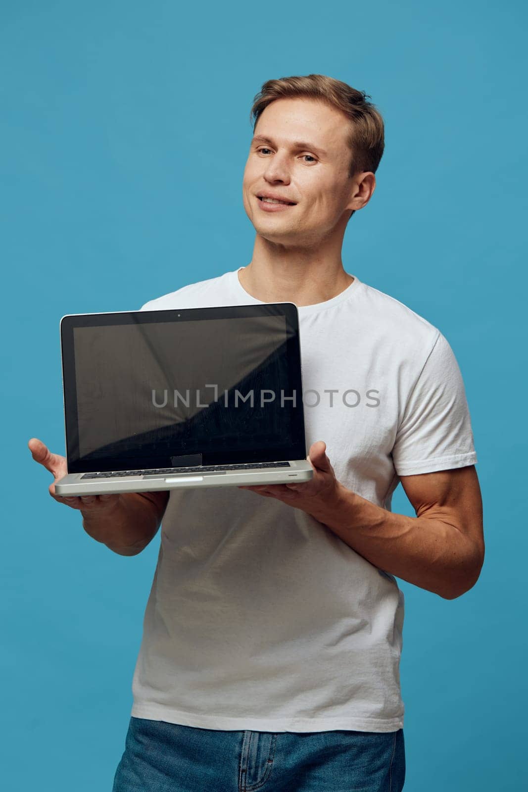 Smiling man presenting open laptop device standing against vibrant blue studio background. Focused on technology and communication, this portrait captures a cheerful professional in a casual outfit. Concept: digital confidence