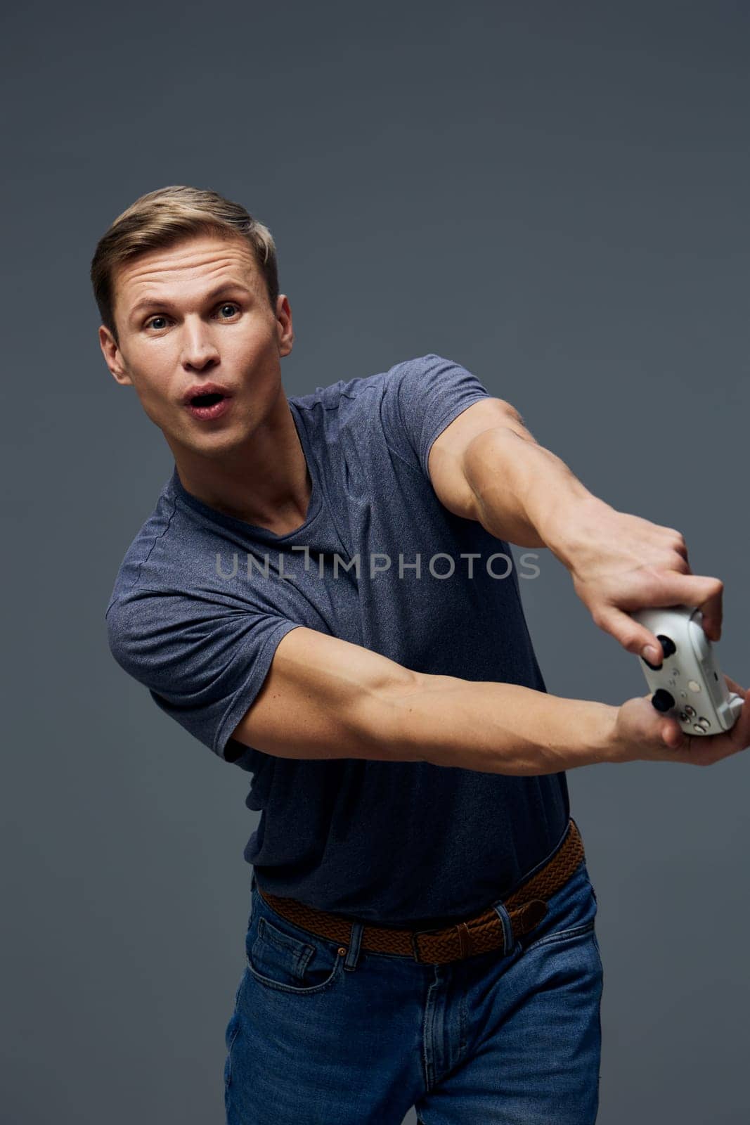 focused young man playing video game holding joystick wearing casual blue t-shirt and jeans with intense facial expression isolated on plain gray background studio portrait digital entertainment concept