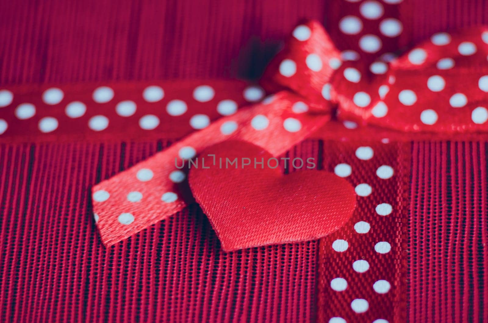 Side view of red tiny heart and gift ribbon with white polka dots tied into a bow, placed on a textured red and pink striped background.
