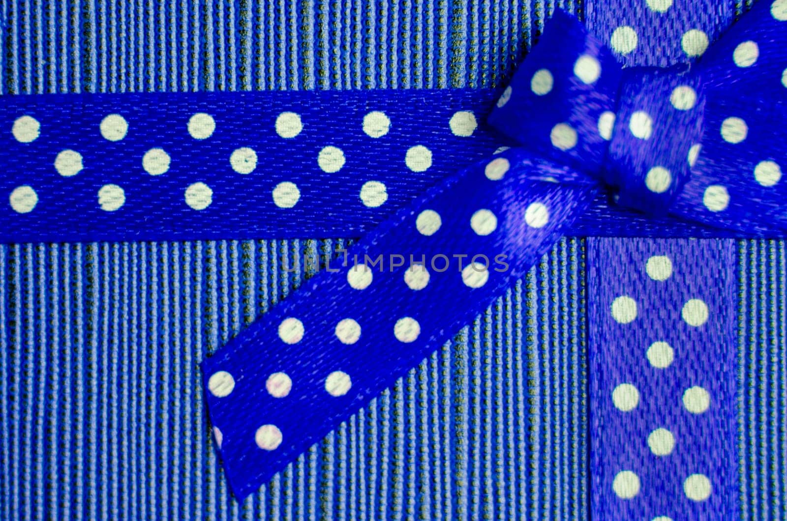Close up of a blue gift ribbon with white polka dots tied into a bow, placed on a textured red and pink striped background.