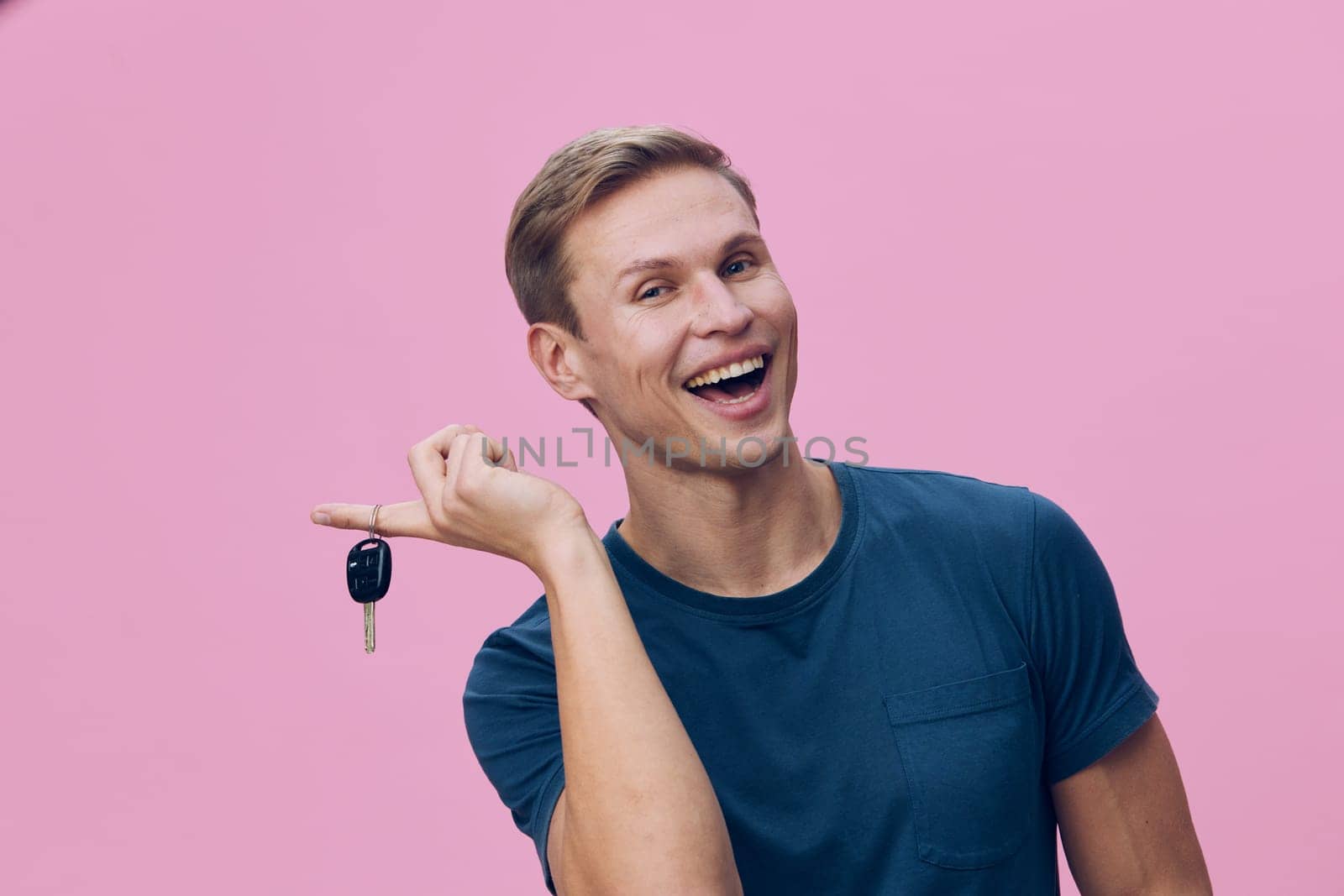 Cheerful man with short light brown hair, dressed in casual blue t-shirt, smiling broadly while holding car keys, standing against a plain pink background. Concept: happiness of new beginnings, personal success, celebration of achievement.