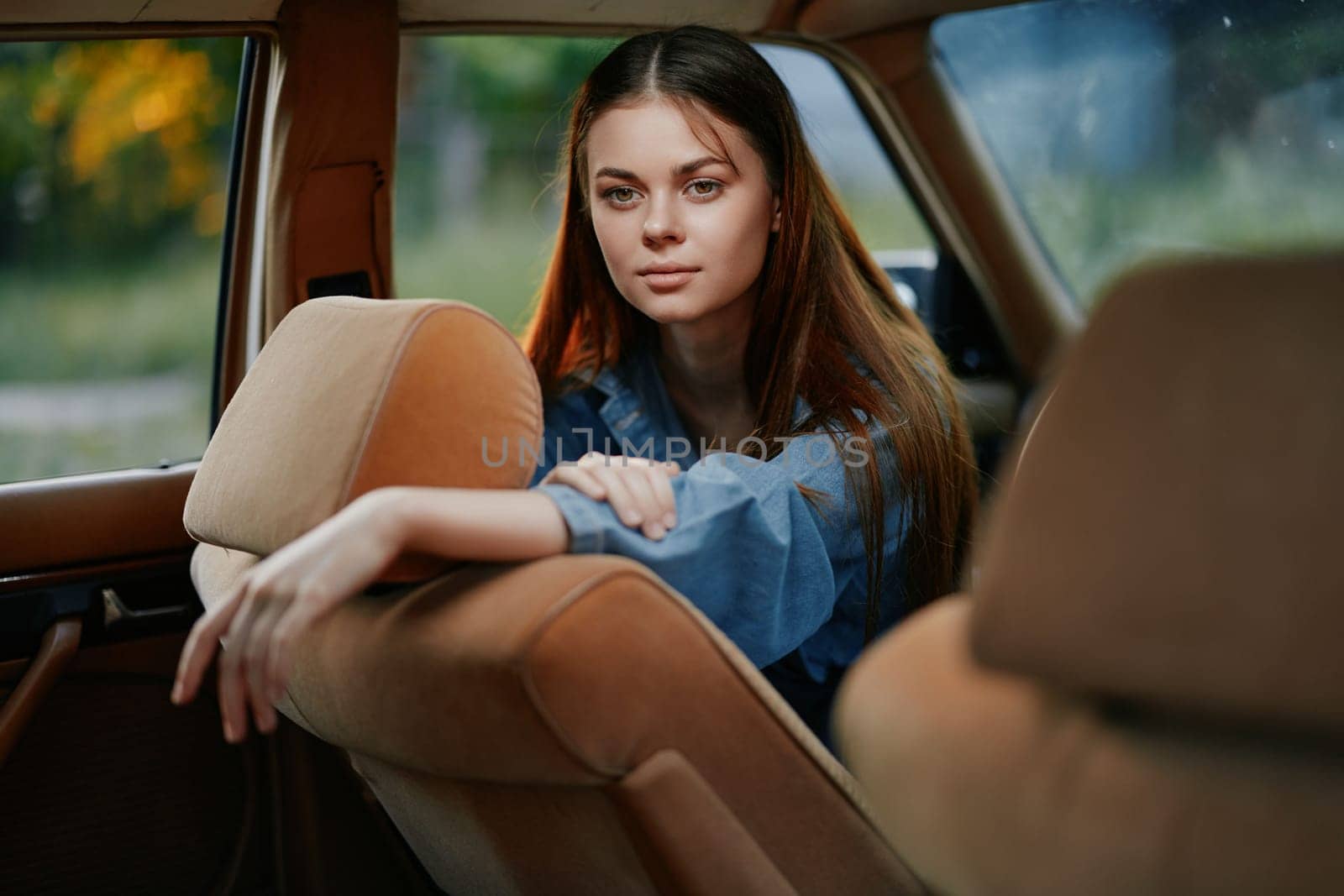 A young woman in a blue shirt sits in the back seat of a vintage car, gazing thoughtfully out the window. The warm colors of the interior contrast with the cool tones of the outdoor scenery, creating