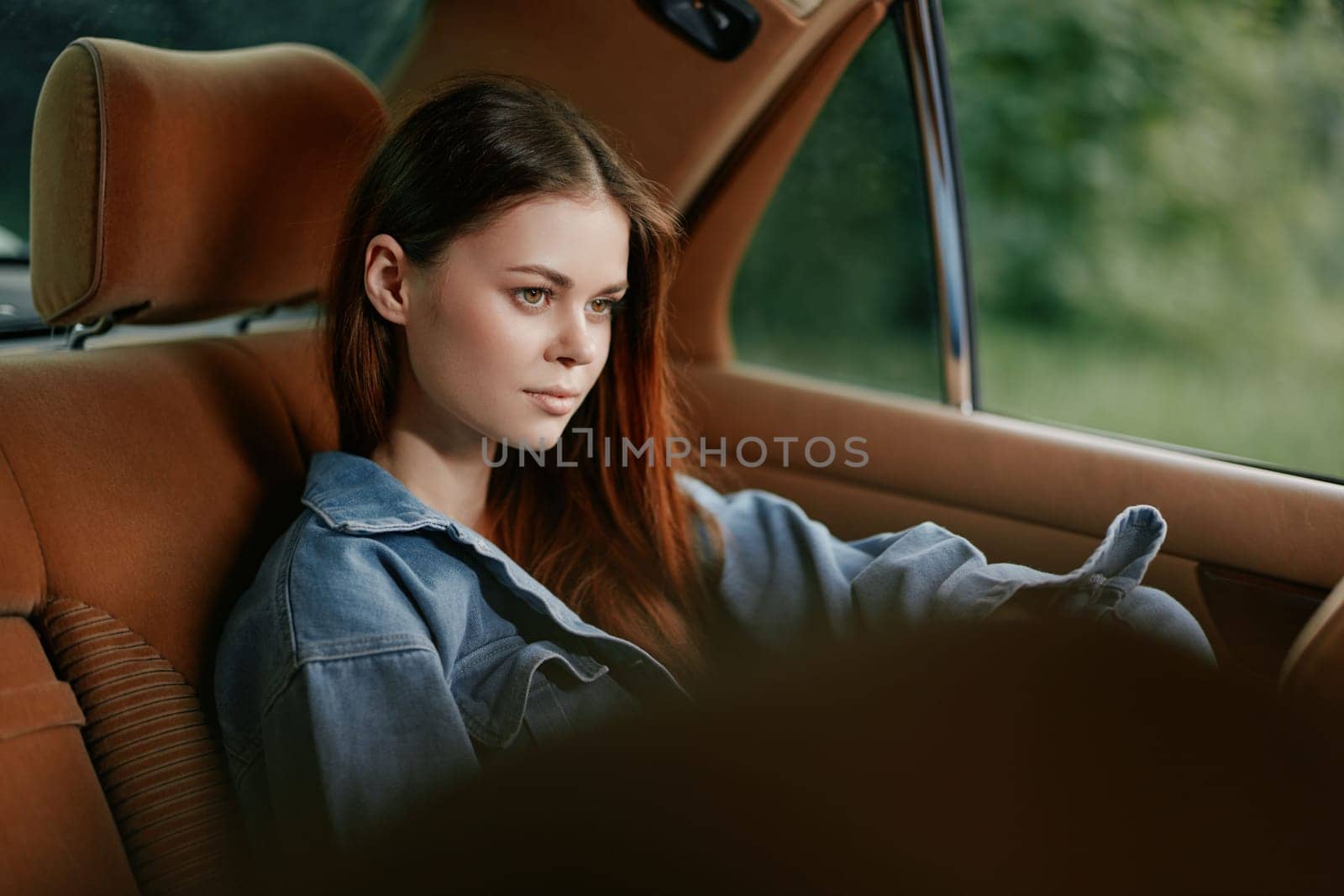 A thoughtful young woman dressed in denim relaxedly sits in a classic car, reflecting while enjoying the lush green scenery outside on a peaceful day.