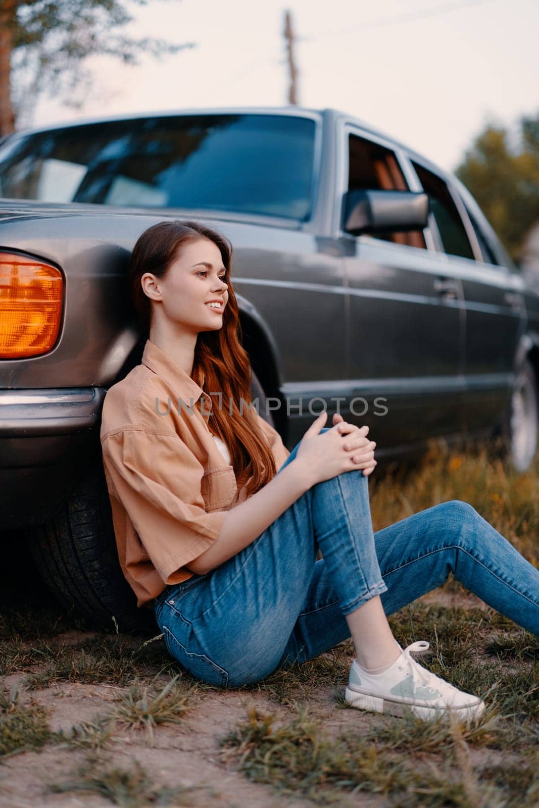 Stylish young woman in casual clothing sitting beside a classic vintage car, embodying a carefree lifestyle in a serene outdoor setting, ideal for lifestyle and automotive themes.