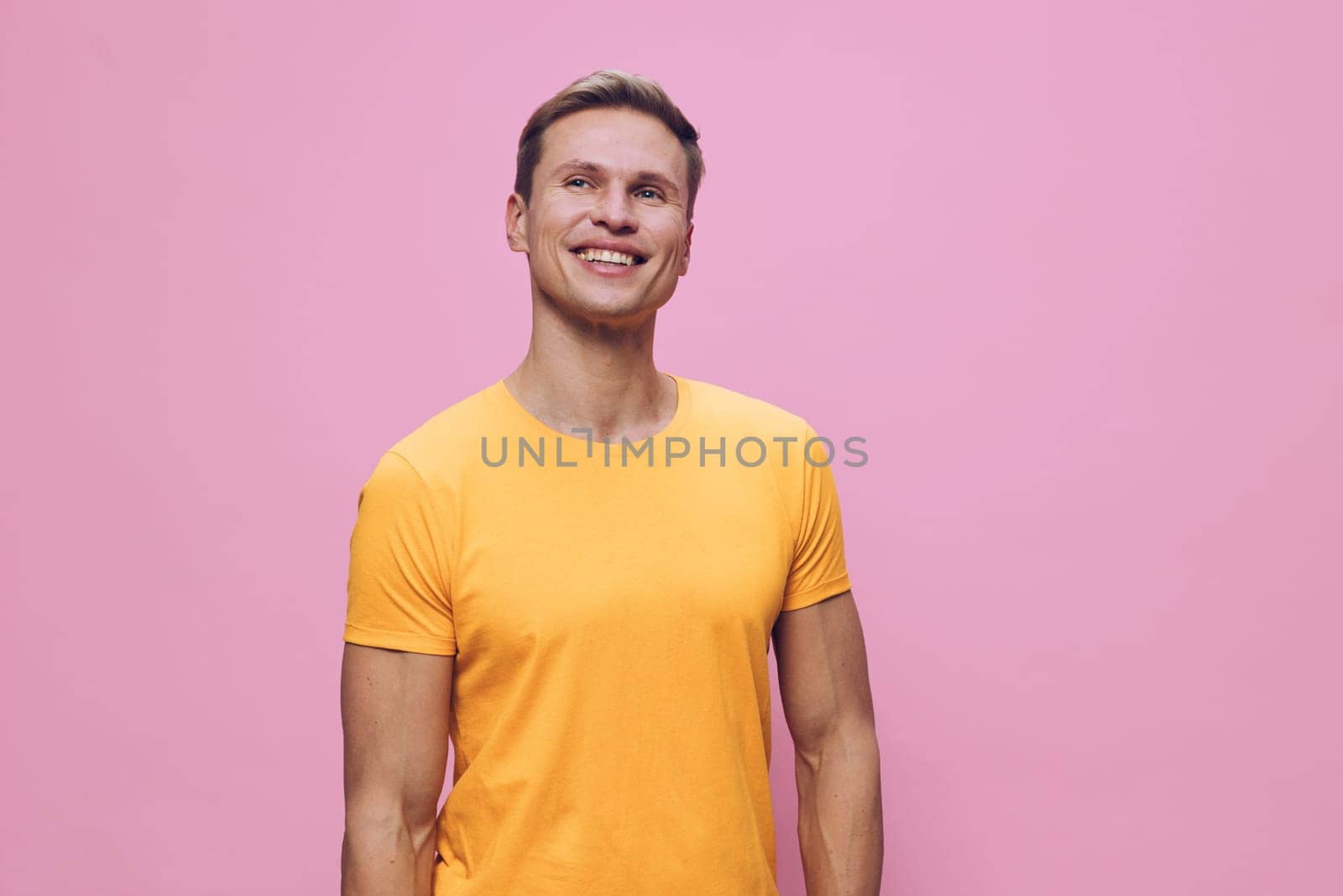Happy man with short brown hair in casual yellow t-shirt smiling broadly while standing against a plain pink backdrop, conveying positivity, friendliness, and warmth, studio photo for lifestyle and health themes. Concept: joy of everyday life.