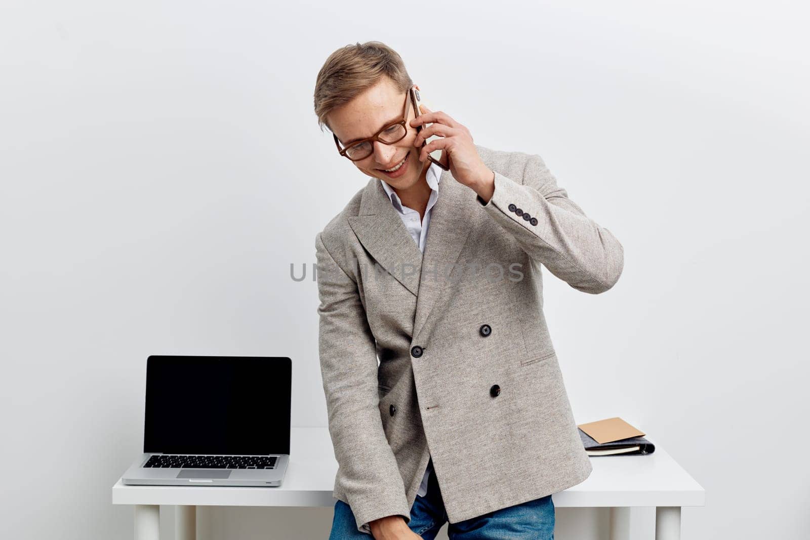 Friendly young man in smart casual clothing engaging in phone conversation at workplace, standing near desk with open laptop and notepad, cheerful expression, modern office environment with minimalist white background, concept: professional communication.