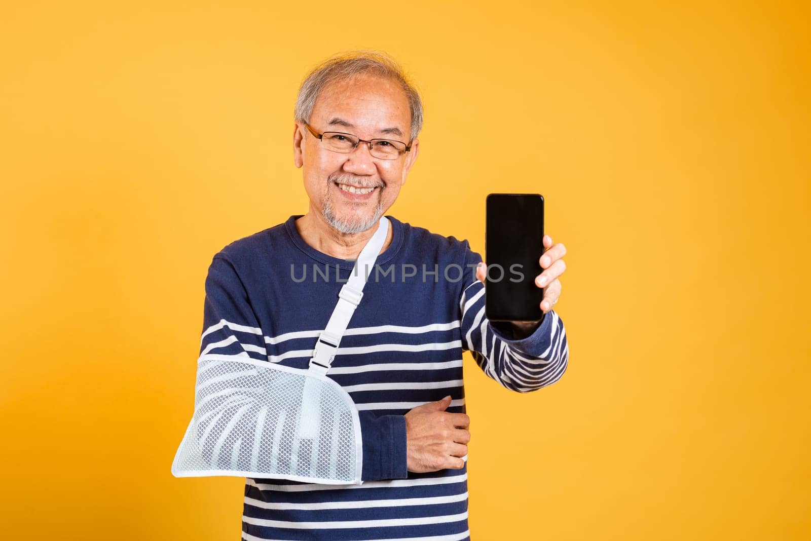 Portrait Asian old man cast his arm and showing mobile phone blank screen studio isolated yellow background, elderly man accident broken arm wear splint treatment, insurance medical care application