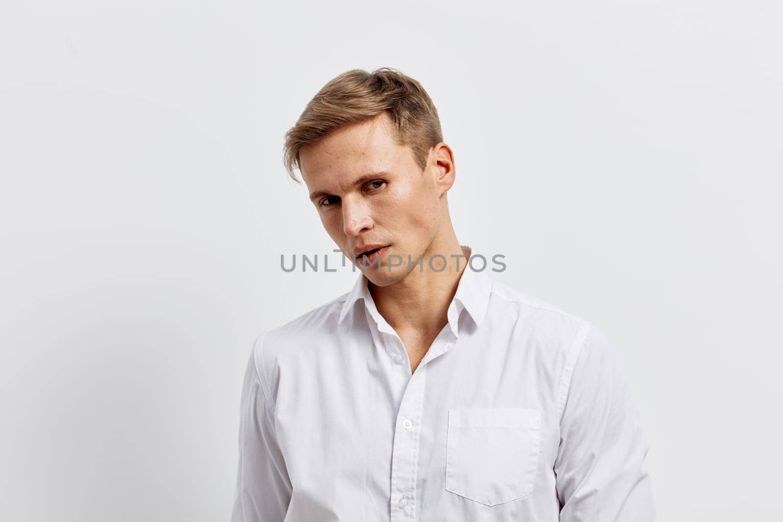 Stylish young man with short hair and fair skin, dressed in a white shirt, posing with a slight tilt, looking at the camera with a serious expression. Background is plain and neutral, emphasizing his confident mood and modern appearance. Concept: professionalism and style.