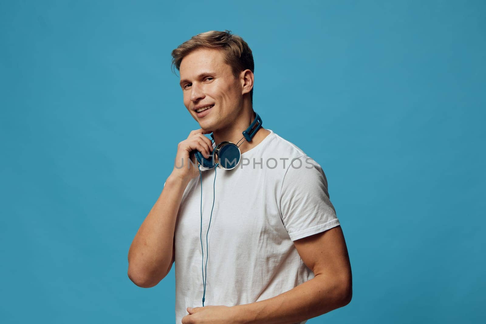 Smiling man with headphones wearing casual white t-shirt stands against a plain blue background, enjoying music. Concept: music enjoyment, leisure, youth, entertainment, relaxed style, studio shot
