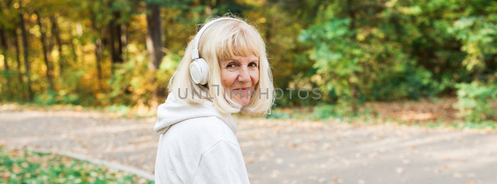 Banner senior woman walking with headphones in the park during fall, copy space. Active lifestyle, happiness, movement, freedom, and mindful aging in nature