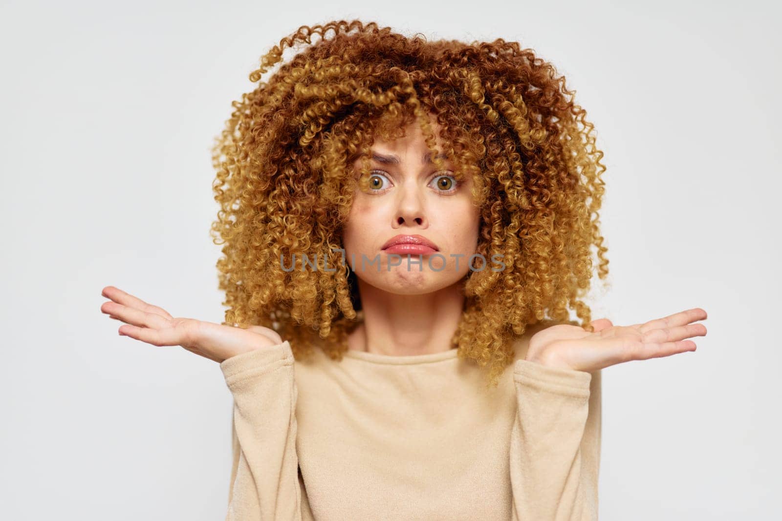 A young woman with voluminous curly hair appears confused, raising her hands in a gesture of uncertainty against a plain gray background The image evokes emotions of inquiry and contemplation, makin