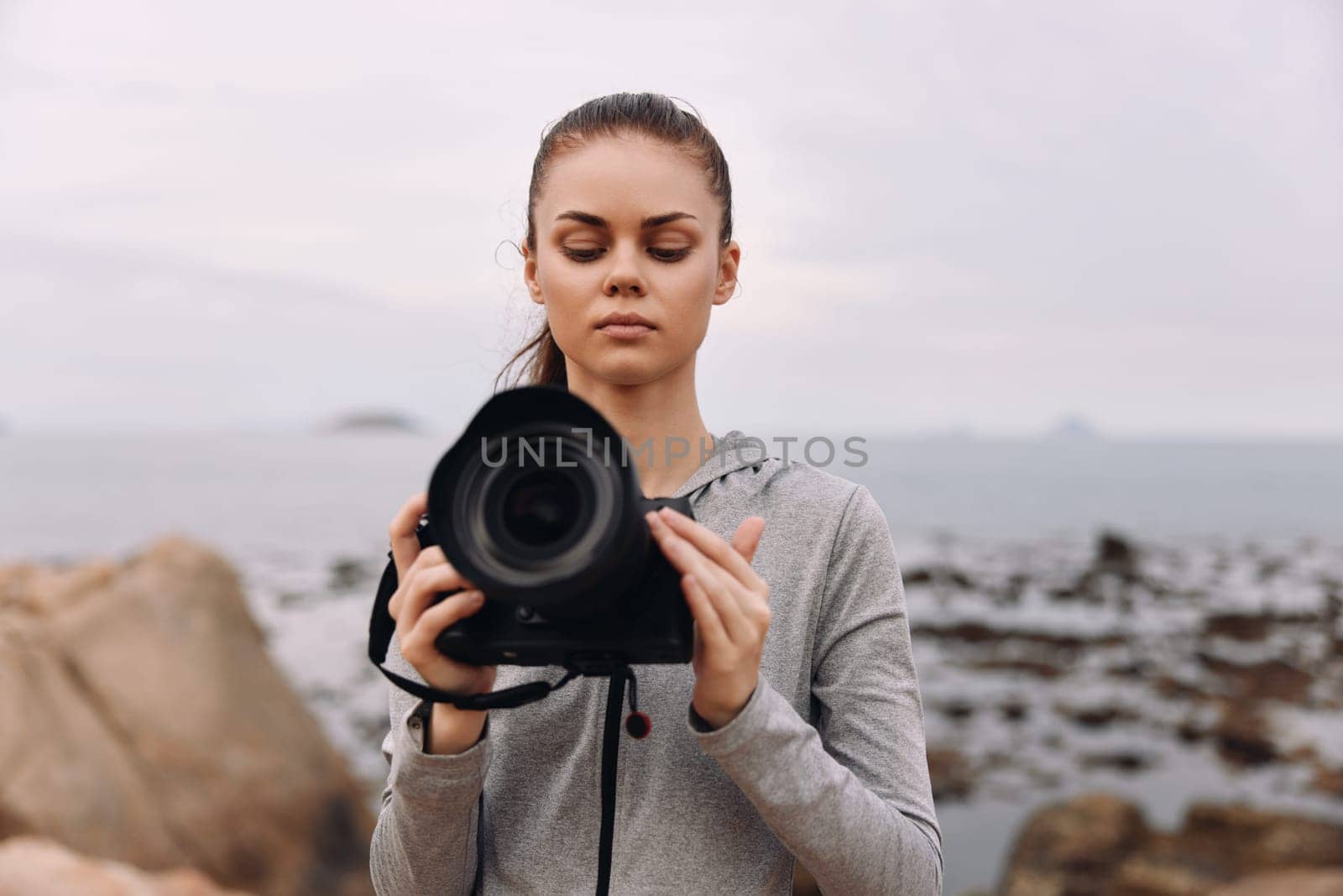 A dedicated young female photographer in a gray hoodie, focused on her camera at a tranquil beach scene with rocky shore under a soft sky, perfect for expressing creativity and nature s beauty