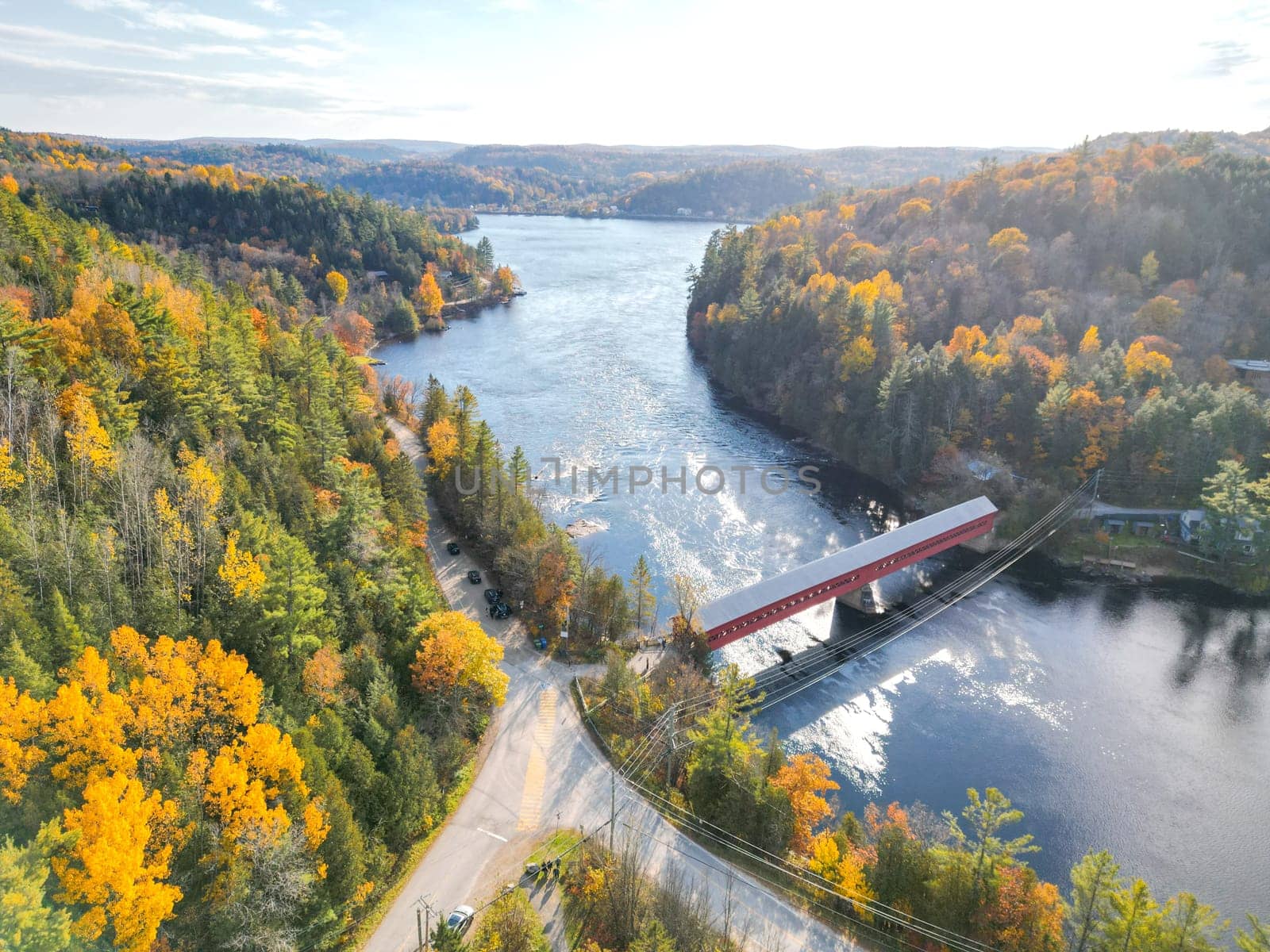 The image shows a small red bridge over a river in a beautiful autumn forest. The trees are in full foliage and the leaves are turning yellow, orange, and red. The water is calm and crystal clear.