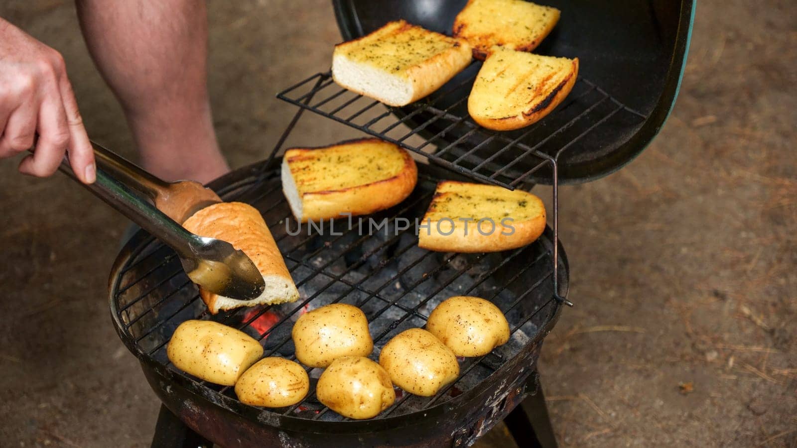 A close-up shot of a charcoal grill with a grate full of potatoes and garlic bread. The potatoes are various sizes and the garlic bread is made with thick slices of bread.