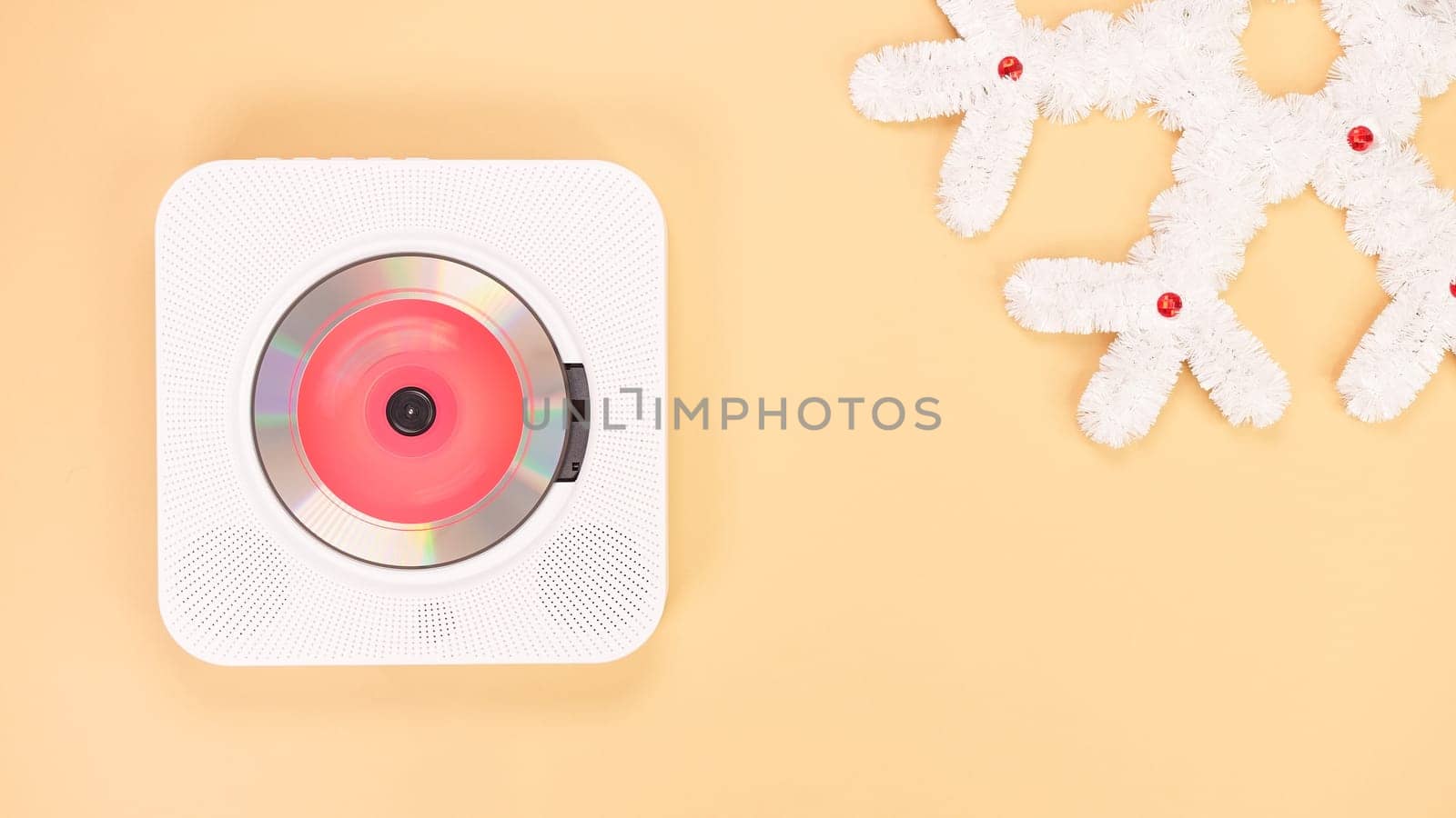 A white portable CD player is positioned on a beige background next to a decorative white snowflake made of tinsel, with a red light shining from inside the CD player.