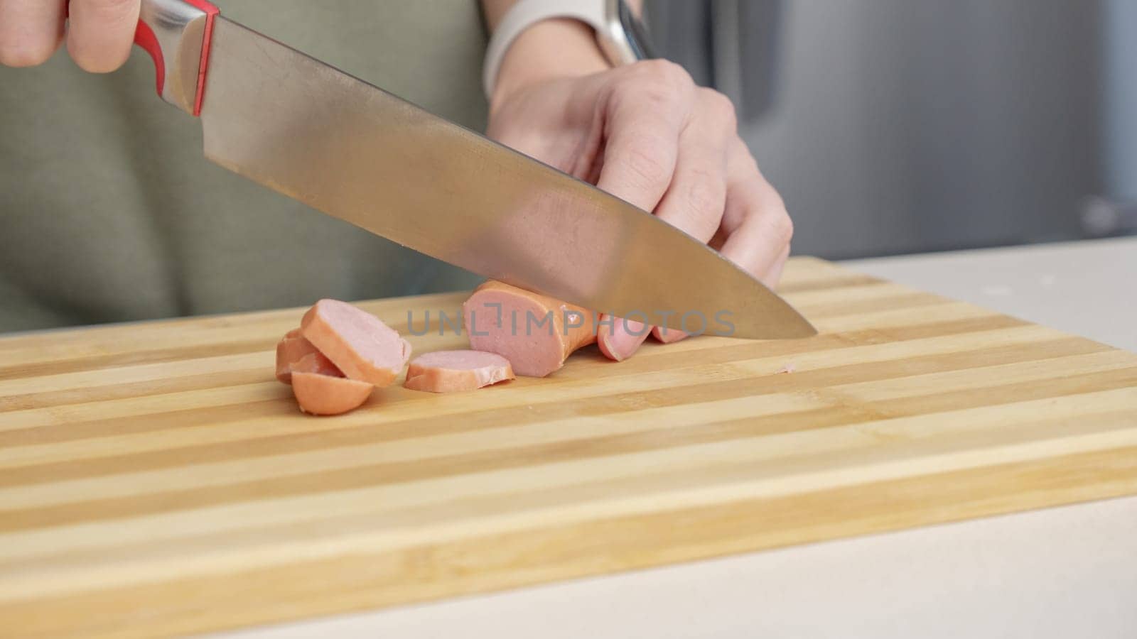 Close-up of a woman cutting sausage on a wooden cutting board in a kitchen setting with different ingredients