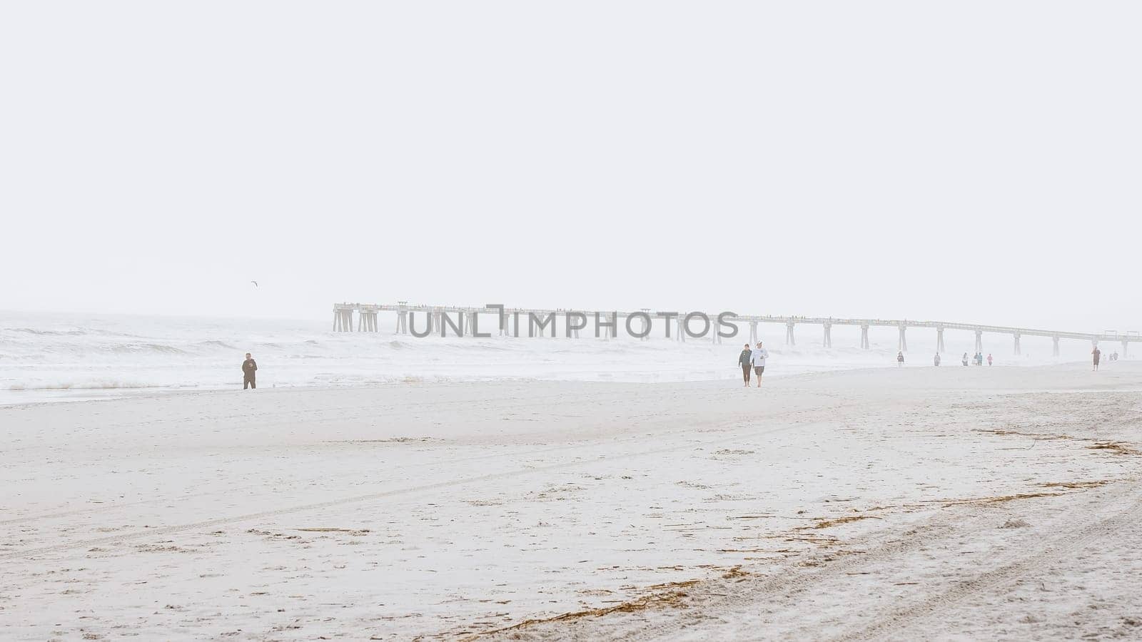 People walking on a beach with a pier in the distance on a foggy day. Jacksonville, Florida USA 03.15.2024