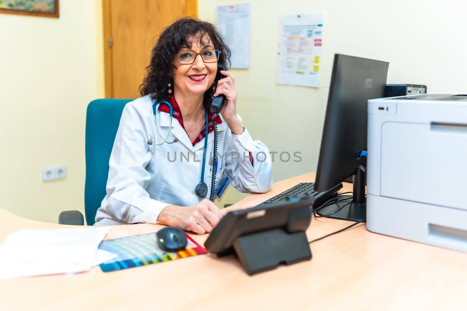 Senior female doctor smiling and answering a phone call, sitting at her desk in a nursing home office