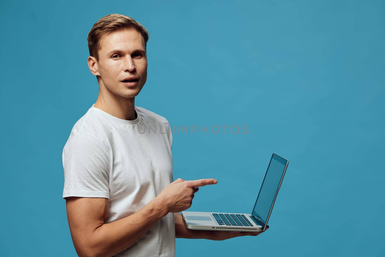 Cheerful young man with short hair wearing a white t-shirt, holding a laptop and pointing at the screen, standing in front of a plain blue background, studio shot, technology, remote work, digital lifestyle concept.