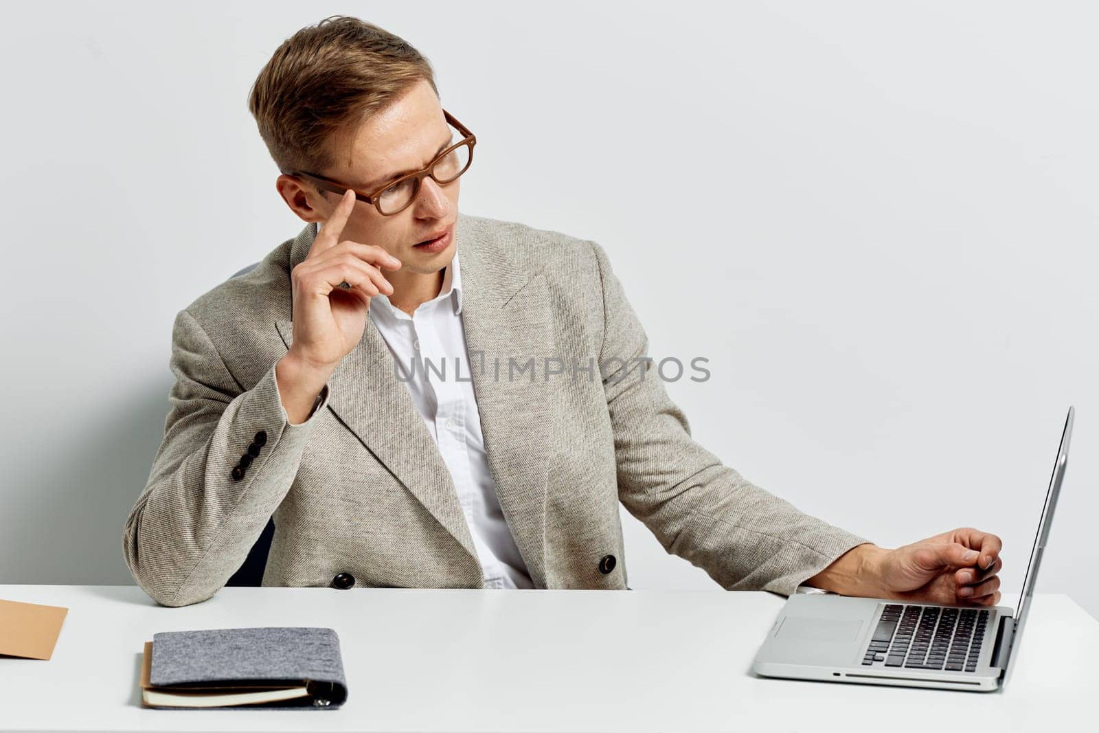 Pensive young businessman with glasses dressed in smart casual attire using laptop and writing notes, isolated on plain white background. Professional, corporate, office work, technology, concentration concept