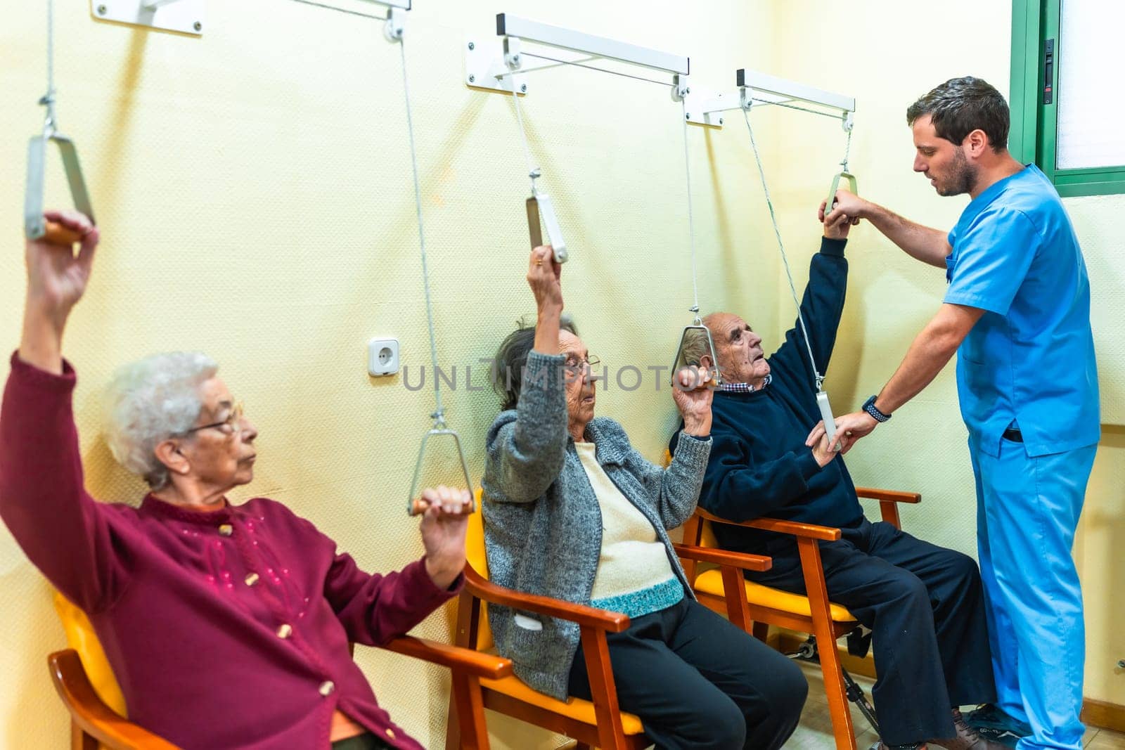 Senior patients practicing physical therapy with pulley system in retirement home, assisted by male nurse