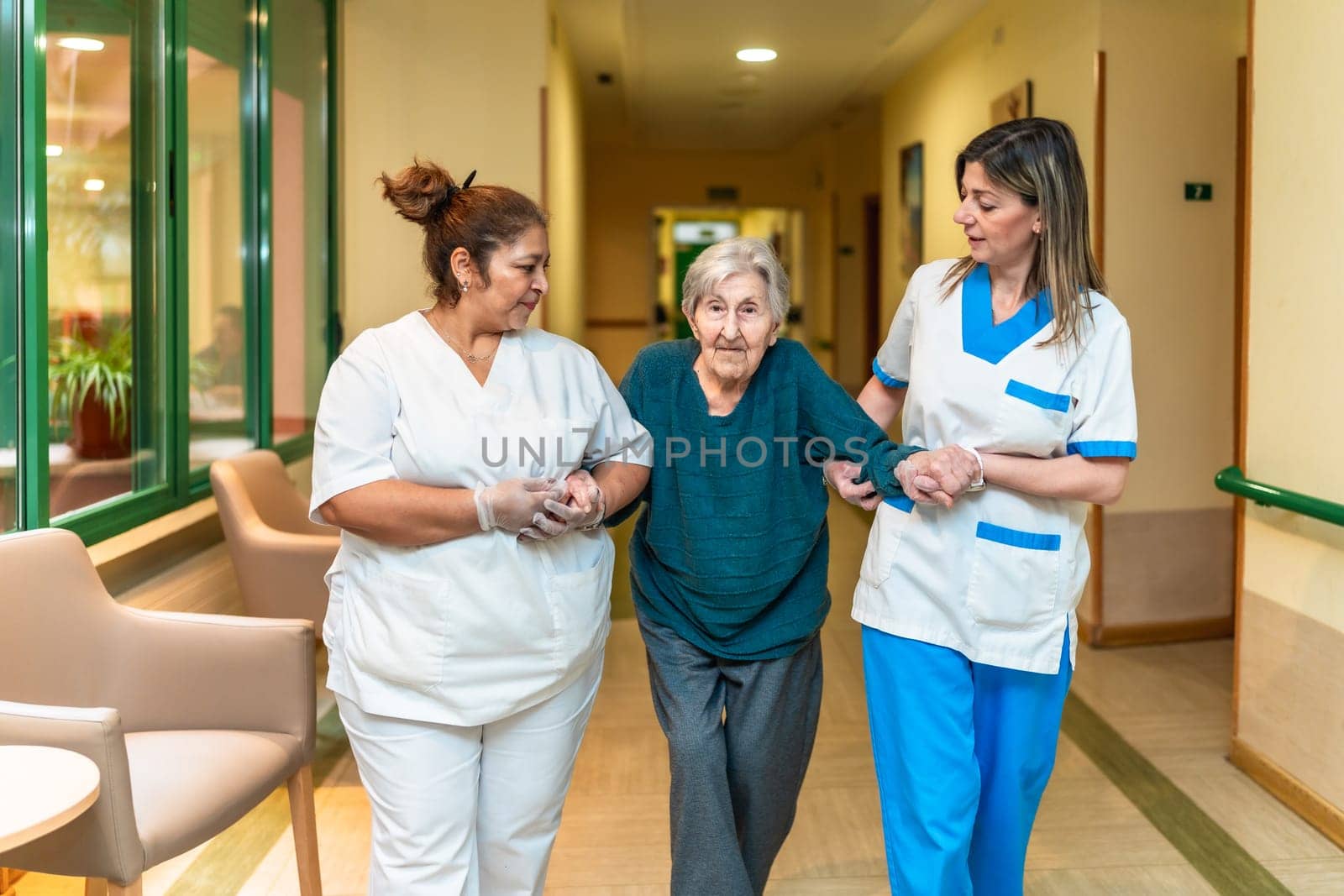Two nurses assisting an elderly woman walking down the corridor of a nursing home, providing support and care