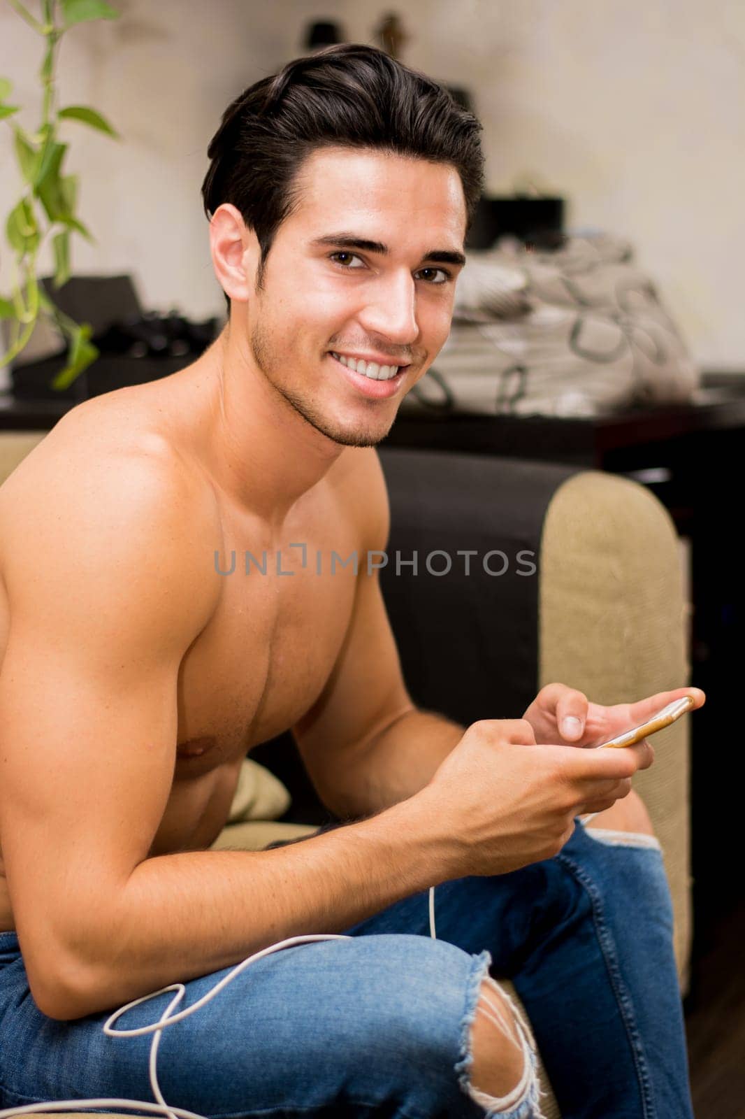 A young man with dark hair sits comfortably on a couch, smiling as he engages with his smartphone. The cozy room has indoor plants in the background, creating a relaxed atmosphere.