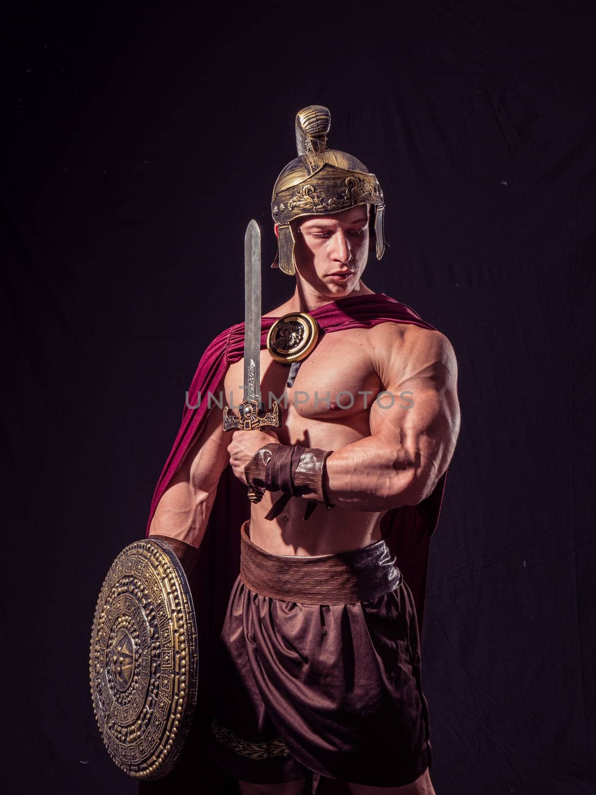 A muscular warrior stands confidently, adorned in traditional armor with a helmet. He grips a sword in one hand and a shield in the other, showcasing strength and readiness.