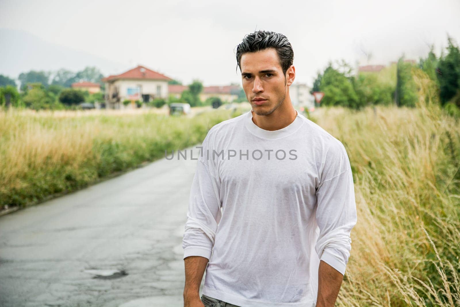 A man stands alone on a quiet rural road, wearing a wet white long-sleeve shirt. The grass around him is tall, and buildings are visible in the background under a cloudy sky.