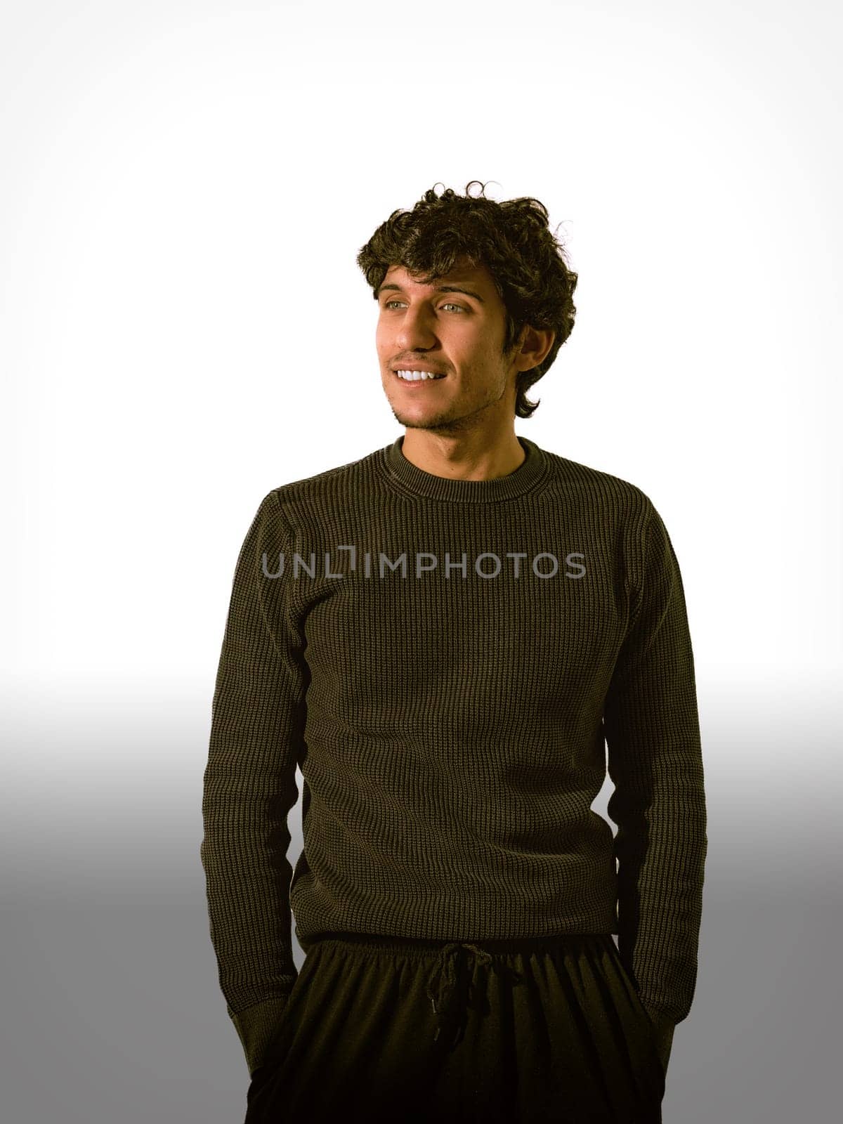 A man with curly hair stands confidently, smiling softly while dressed in a dark sweater. Neutral tones in the background emphasize his relaxed demeanor in a casual indoor setting.