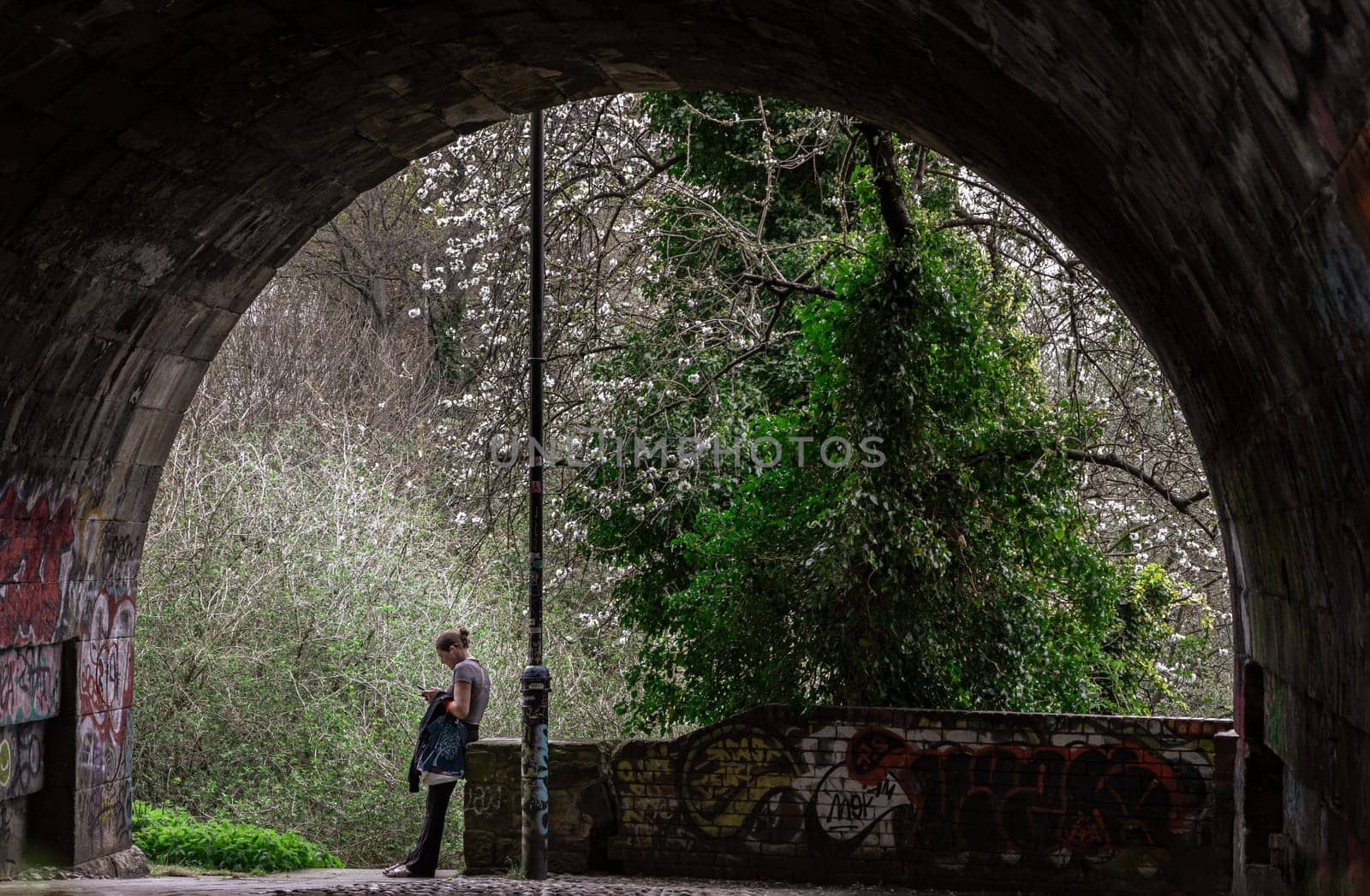 Newcastle, UK - Apr 12, 2025 - View of woman in t-shirt and jeans leans against brick railing wall and looking at a message on her smartphone under the high level bridge at afternoon sunlight. use it as your Wallpaper, Poster and Copy space, Selective focus.