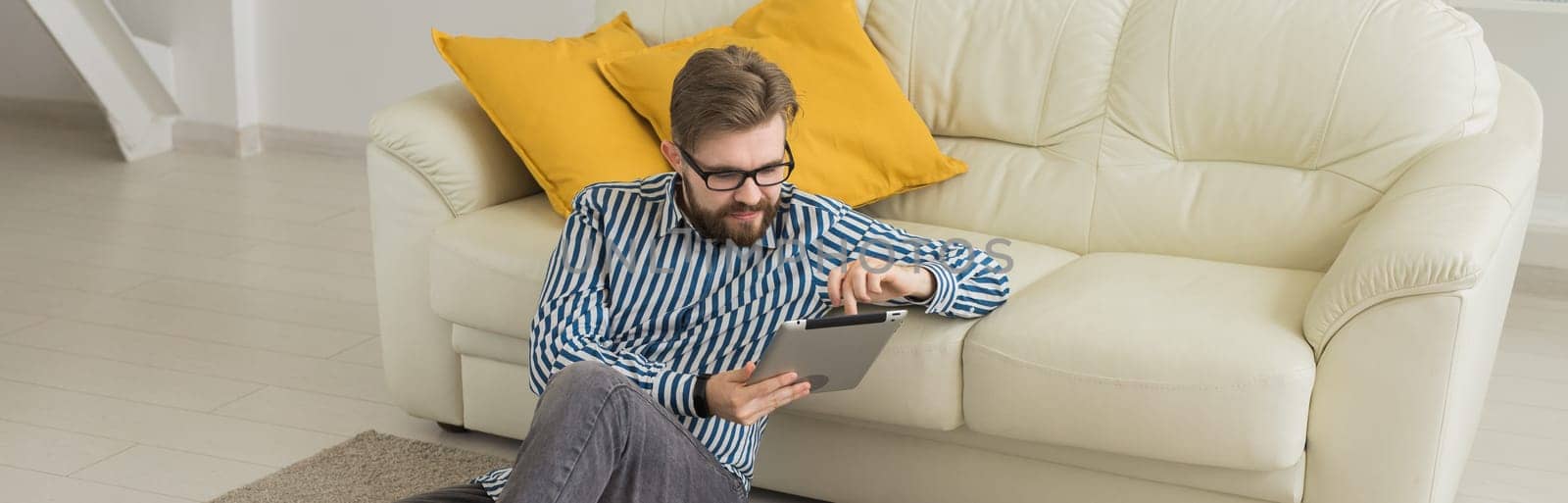 Young man lying on floor and using tablet device near sofa. Digital communication and casual home lifestyle concept. Banner copy space.