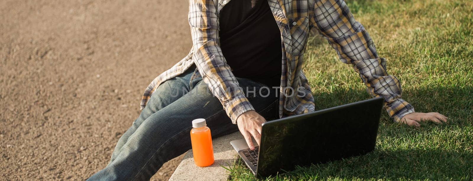 Man working outdoors on a laptop with juice beside him. Concept of remote work, productivity, and casual freelance lifestyle in urban parks. Banner with copy space