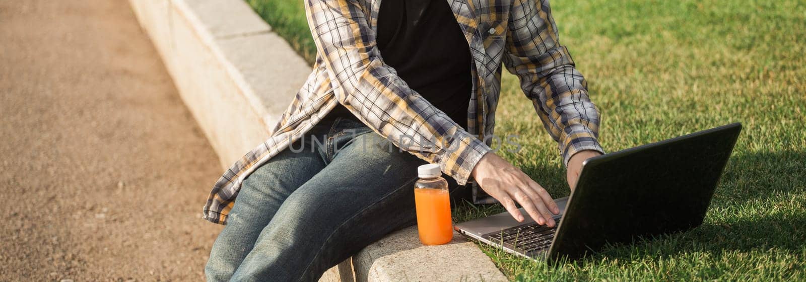 Man working outdoors on a laptop with juice beside him. Concept of remote work, productivity, and casual freelance lifestyle in urban parks. Banner with copy space