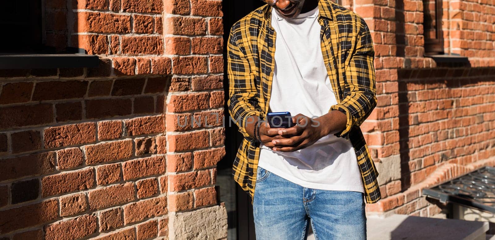 African american man in a plaid shirt texting on smartphone outdoors. Concept of digital communication, connection and casual lifestyle.