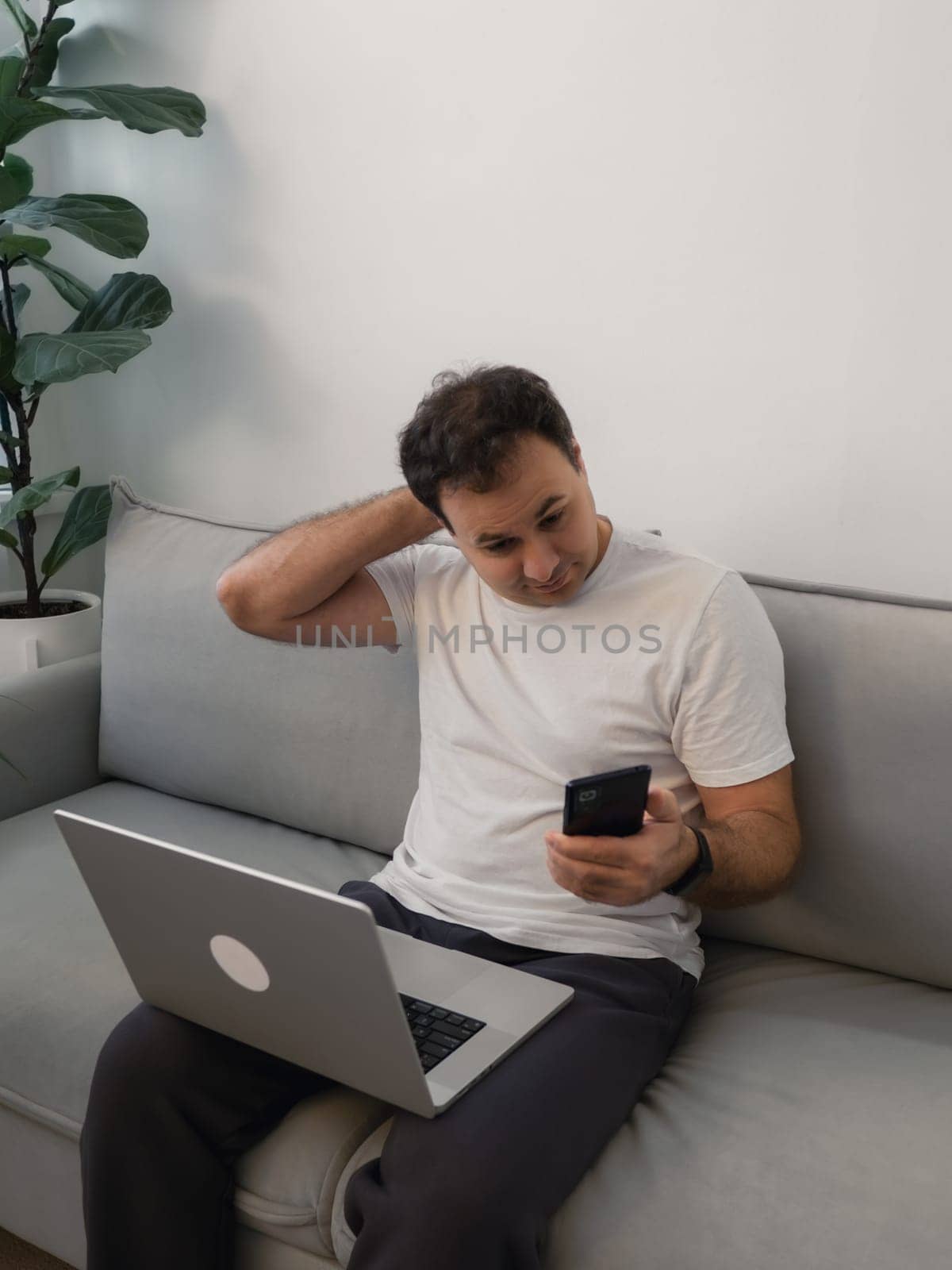 Casual man multitasking with smartphone and laptop showing thoughtful expression. Highlights themes of digital communication, decision making, adapting to modern technology challenges, problem solving