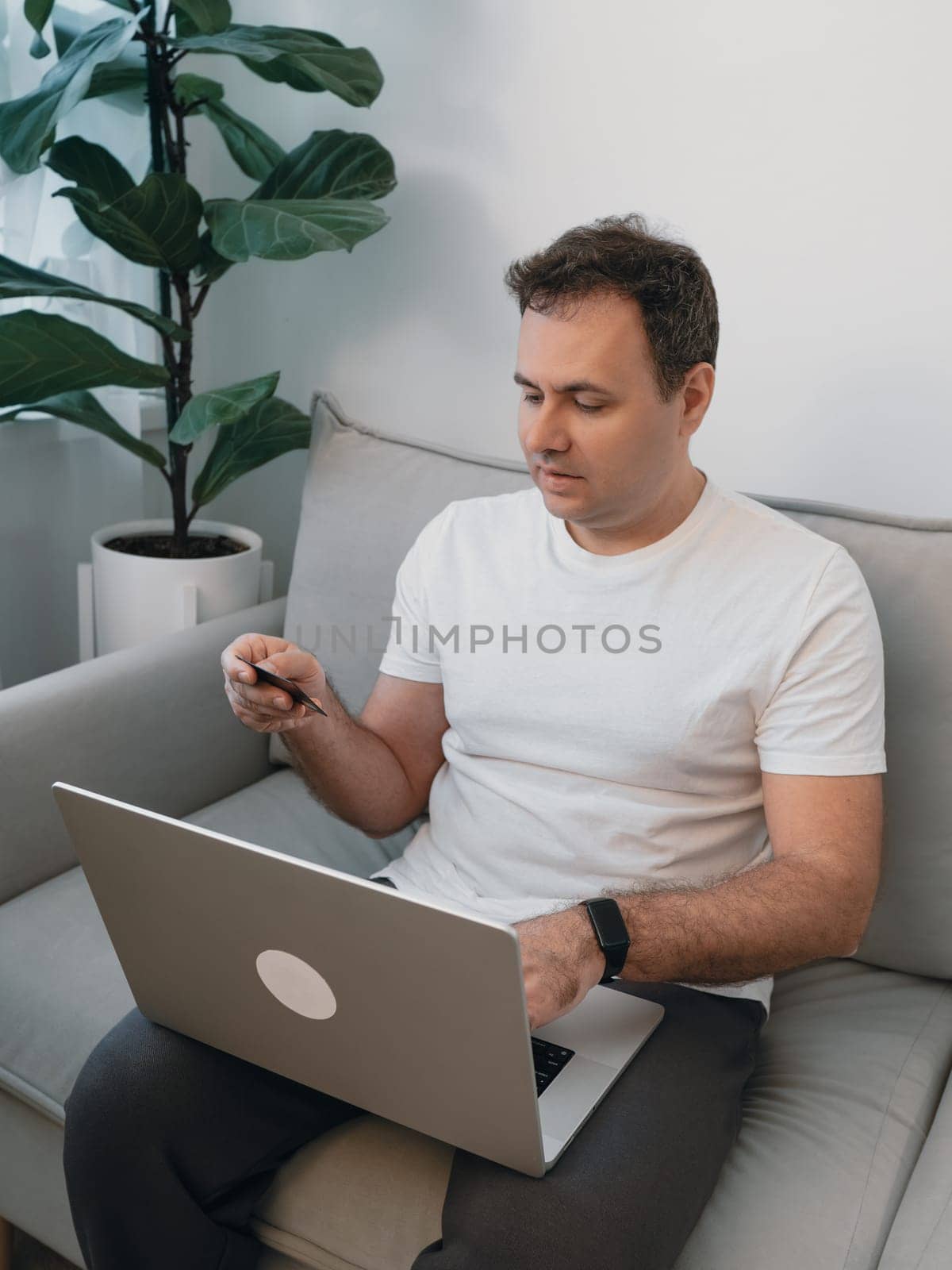 Side view of a man holding a bank card and verifying details in a modern living room with a fiddle leaf fig plant. Concept of secure online payment, digital banking, e-commerce and online shopping