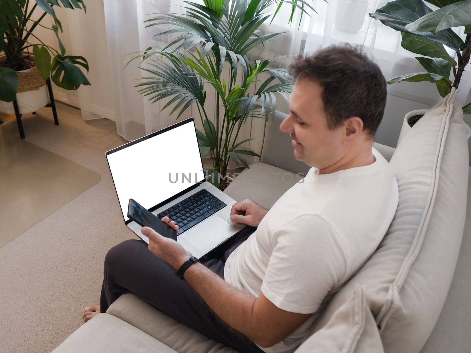 Smiling man sitting with laptop on knees, looking at smartphone. Laptop screen is visible, ideal for mockup use. Concept of positive digital interaction, remote work, and modern technology lifestyle.