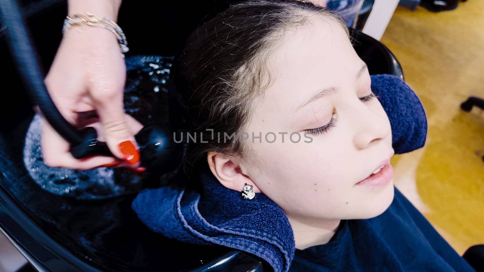 Client relaxes while having her hair washed in a salon basin, the stylist's hands gently massaging her scalp. Water ripples in the basin reflect the care of the salon experience, as a protective cape drapes over the client, ensuring comfort and neatness.