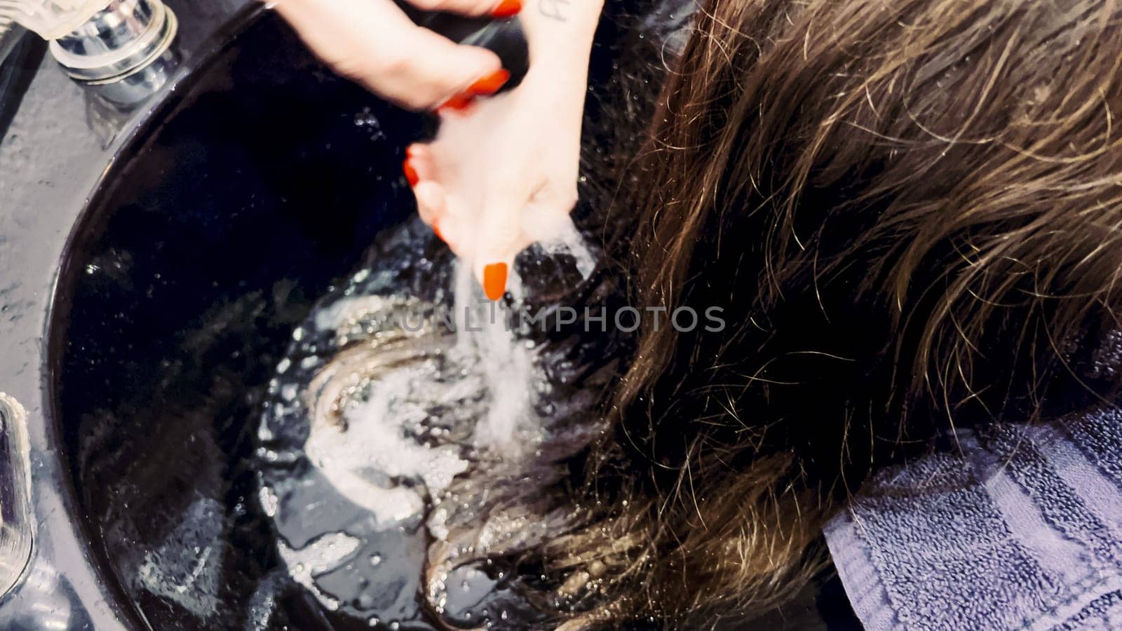 Client relaxes while having her hair washed in a salon basin, the stylist's hands gently massaging her scalp. Water ripples in the basin reflect the care of the salon experience, as a protective cape drapes over the client, ensuring comfort and neatness.